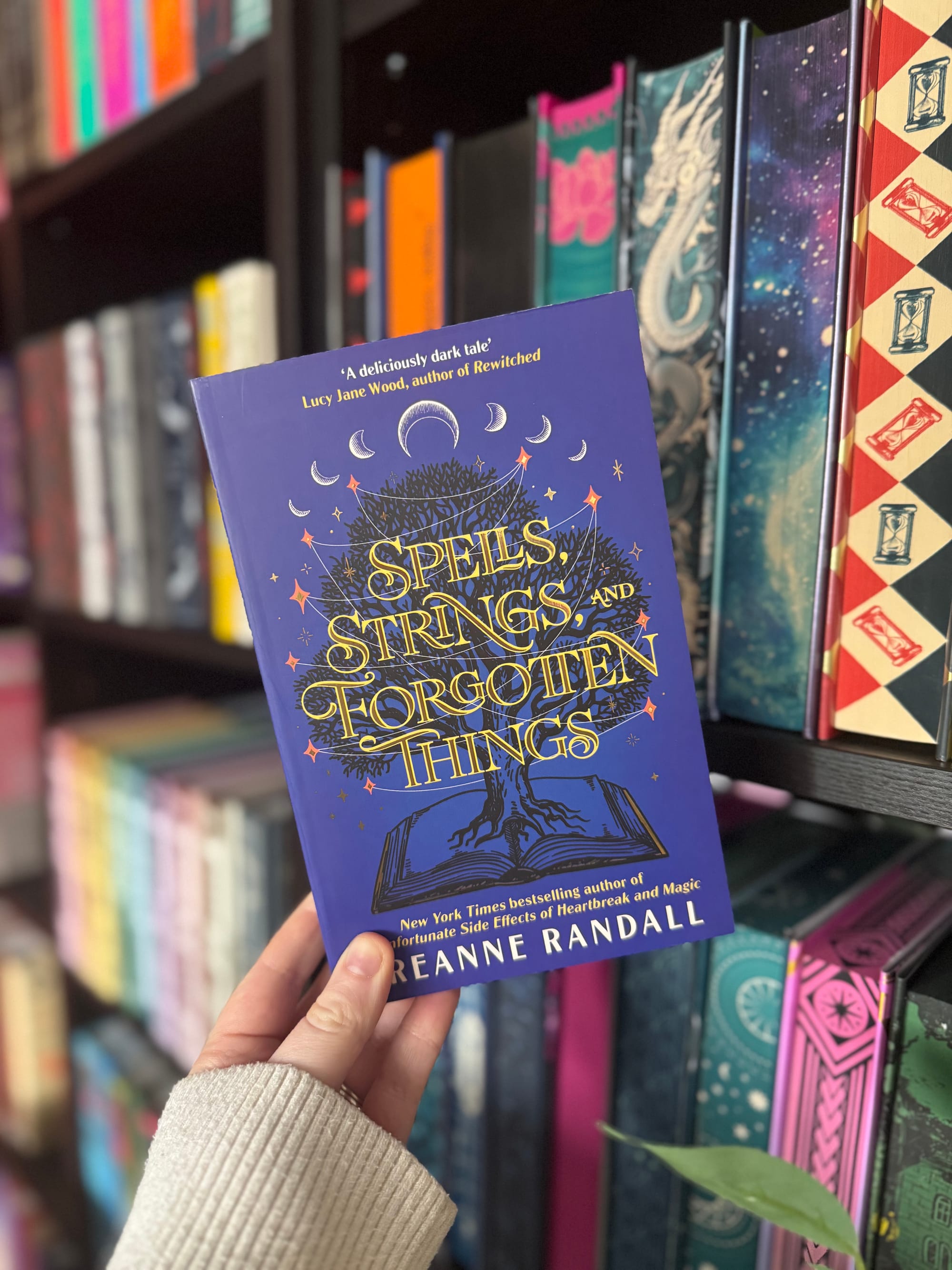 Hand holding a blue book titled "Spells, Strings, and Forgotten Things" in a bookstore. Shelves with colorful books form the background.