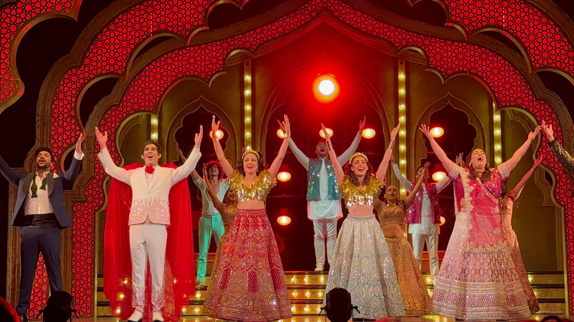Performers in colorful traditional attire dance on a stage with a vibrant red arch backdrop, illuminated by warm spotlights, before an audience.