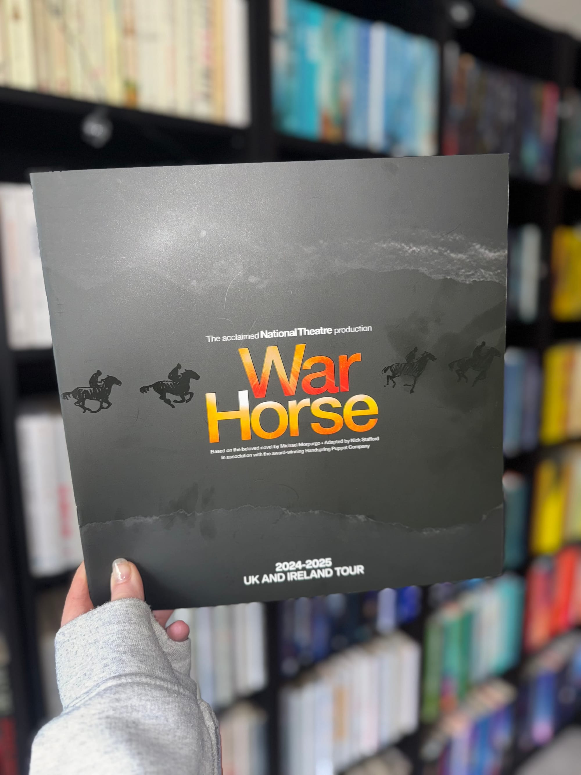 A hand holds a War Horse theatre programme against a blurred background of bookshelves. The programme is black with the title "War Horse" in bold orange letters, featuring silhouettes of galloping horses and riders.