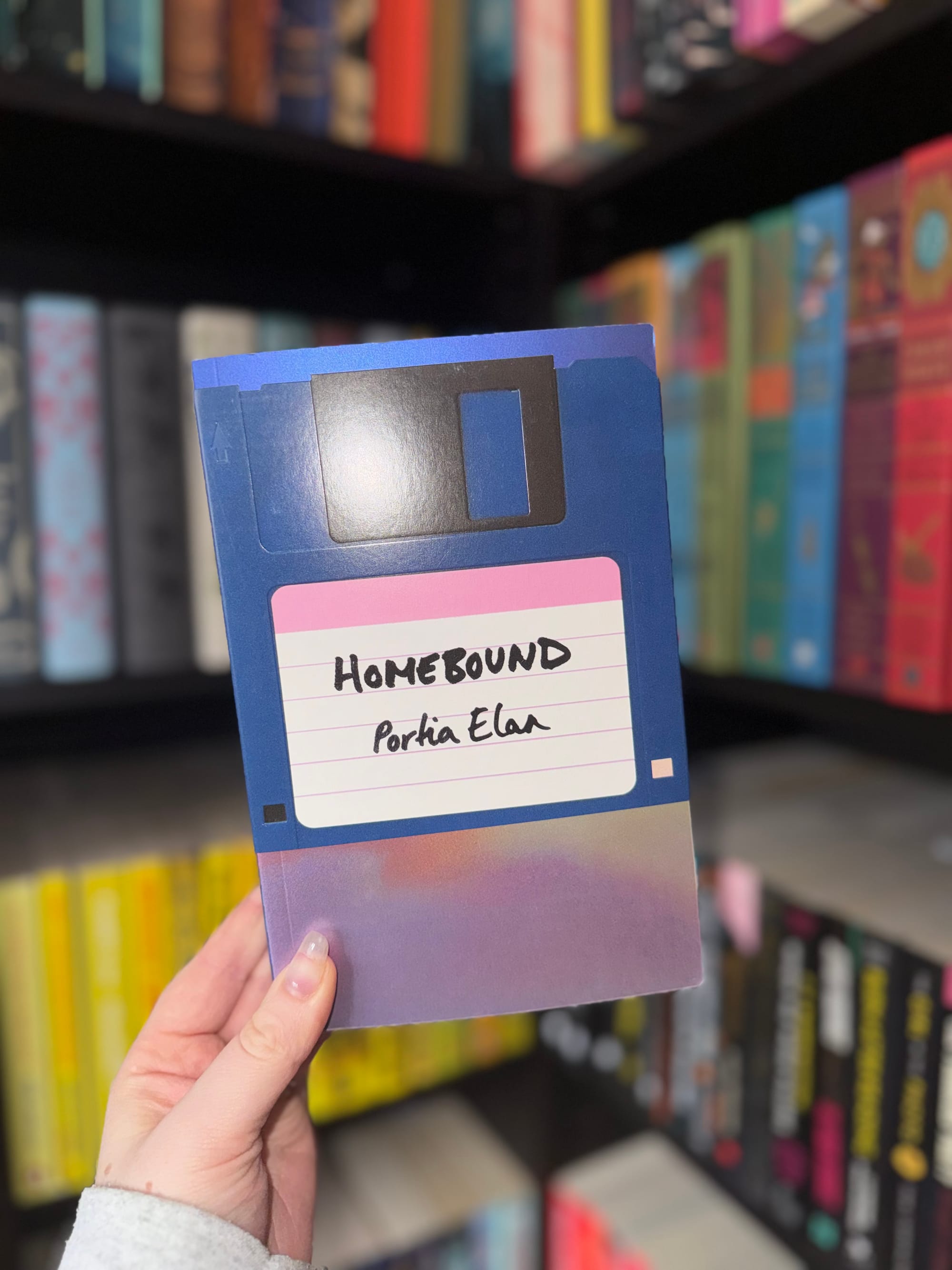 Hand holds a book titled "Homebound" by Portia Elan. Cover has a floppy disk design. Background shows colorful books on shelves.