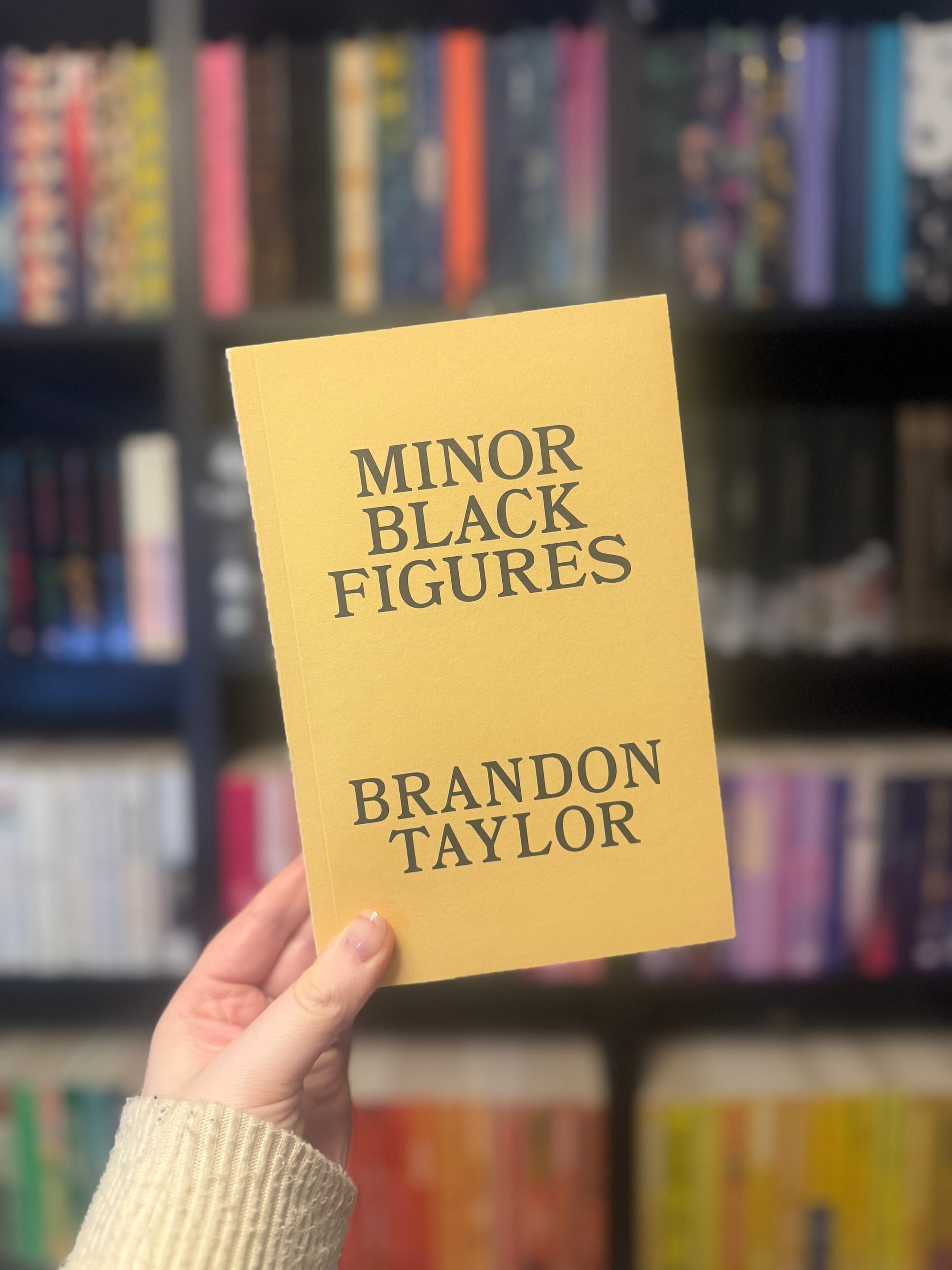Hand holding a yellow book titled "Minor Black Figures" by Brandon Taylor, in a colorful bookstore setting.