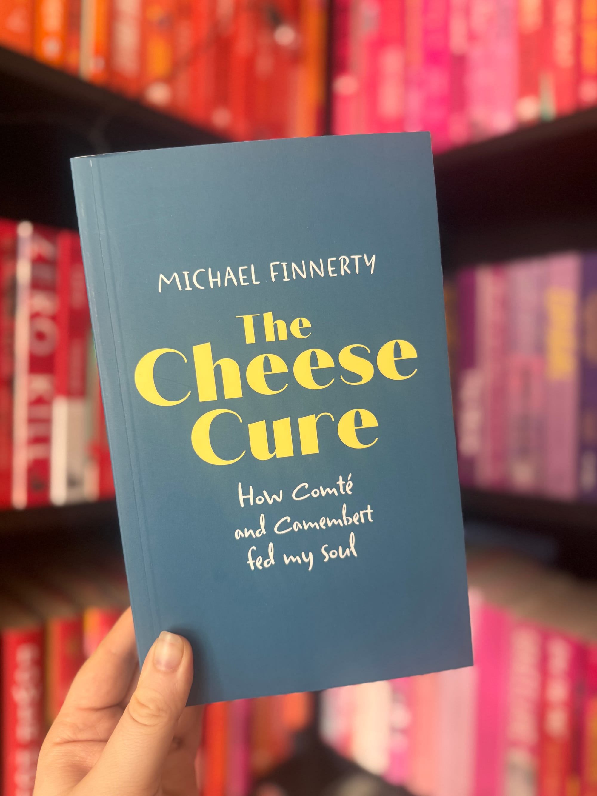 A hand holds a blue book titled "The Cheese Cure" in front of colorful book shelves. The mood is vibrant and literary.