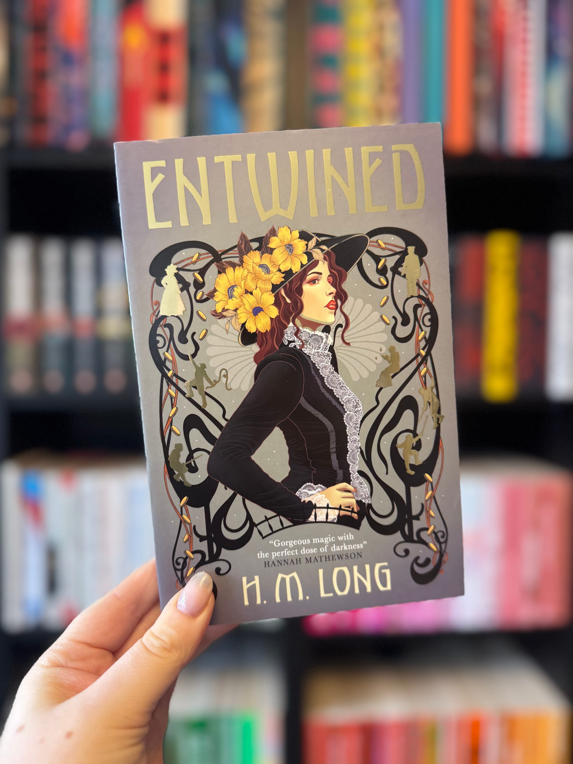 Hand holding the book "Entwined" by H.M. Long, featuring an illustrated woman with flowers in her hair. Blurred colorful bookshelves behind.