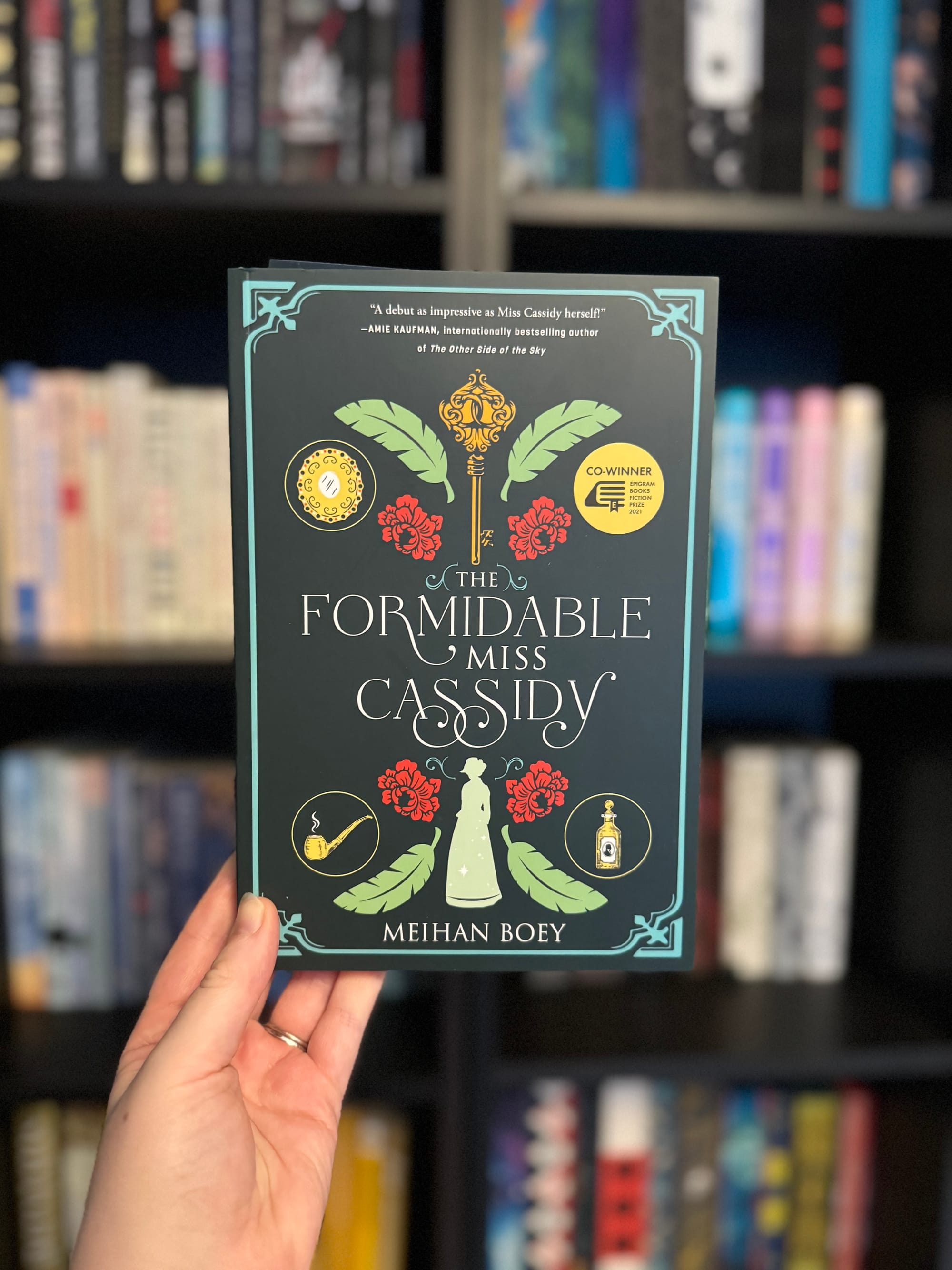 Hand holding "The Formidable Miss Cassidy" book by Meihan Boey. Green and gold cover with floral design. Bookshelf blurred in background.