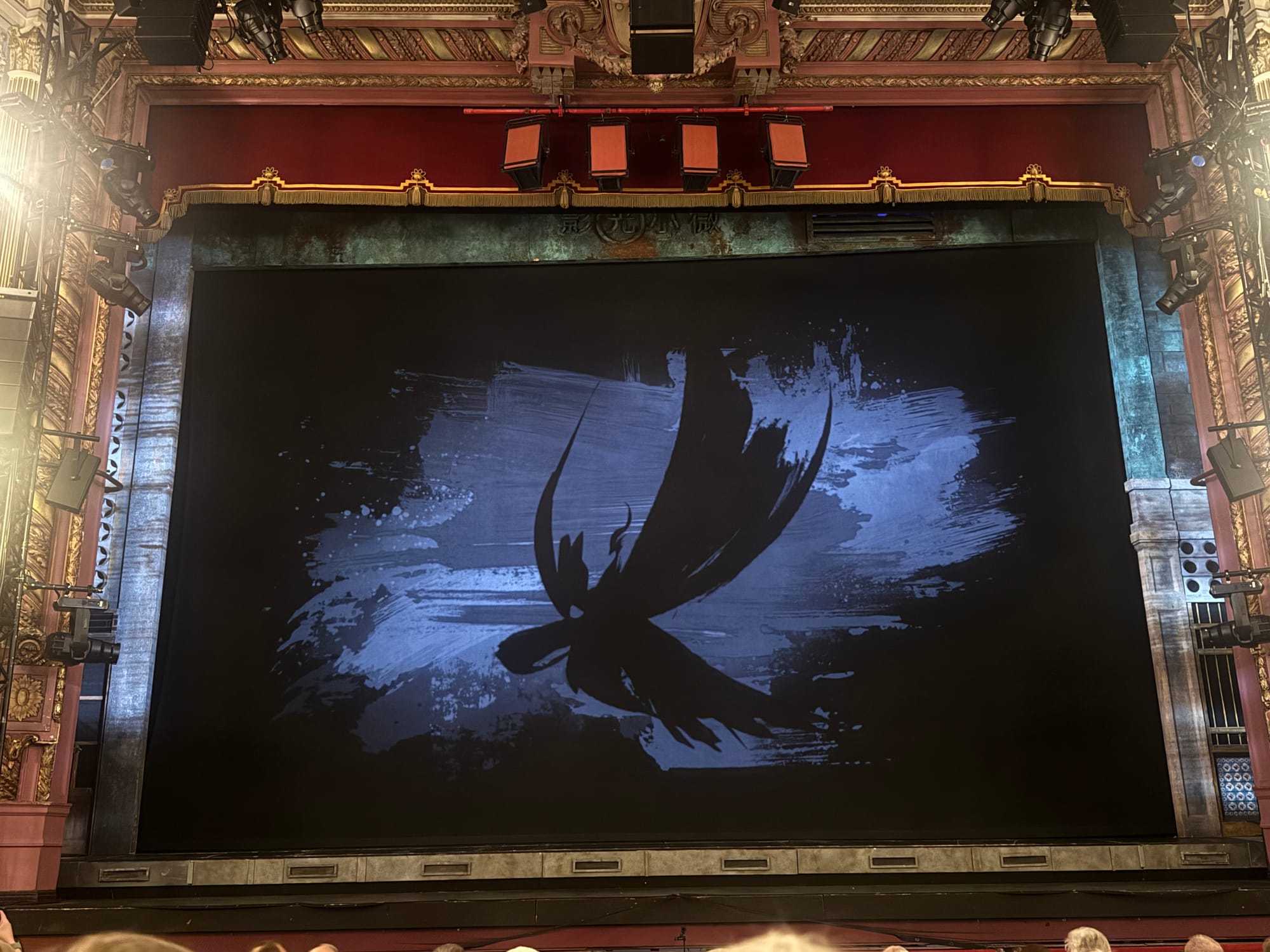 Stage with ornate red and gold decor, featuring a large black and blue curtain with an abstract bird design. Dim lighting, audience visible.