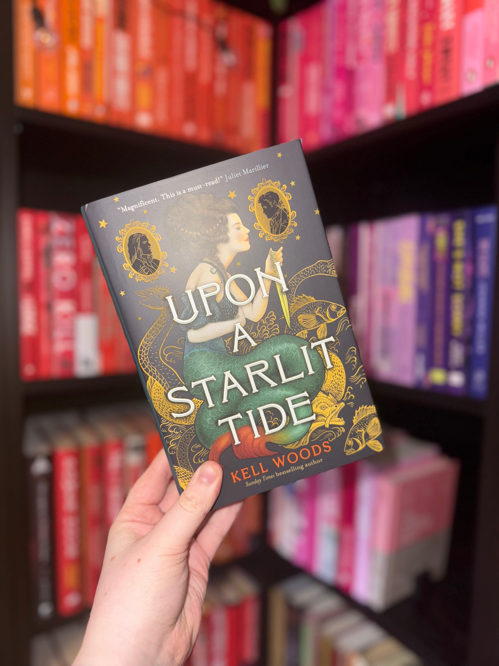 Hand holding "Upon a Starlit Tide" book by Kell Woods. Colorful bookshelf backdrop; red, pink, blue spines visible. Book cover features ornate design.