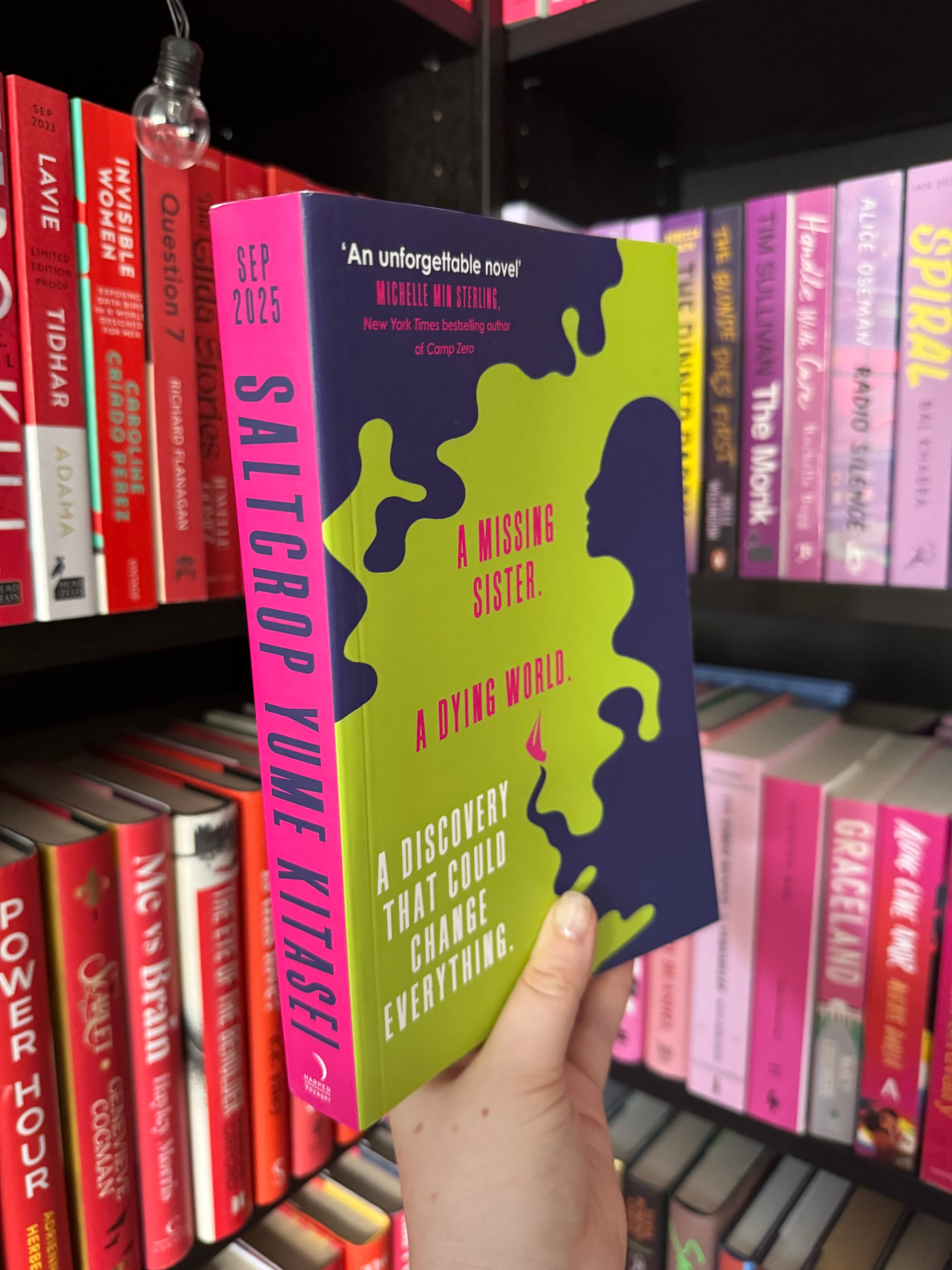 Hand holding a colorful book titled "Saltcrop." Text: "A Missing Sister. A Dying World. A Discovery That Could Change Everything." Bookshelf background.