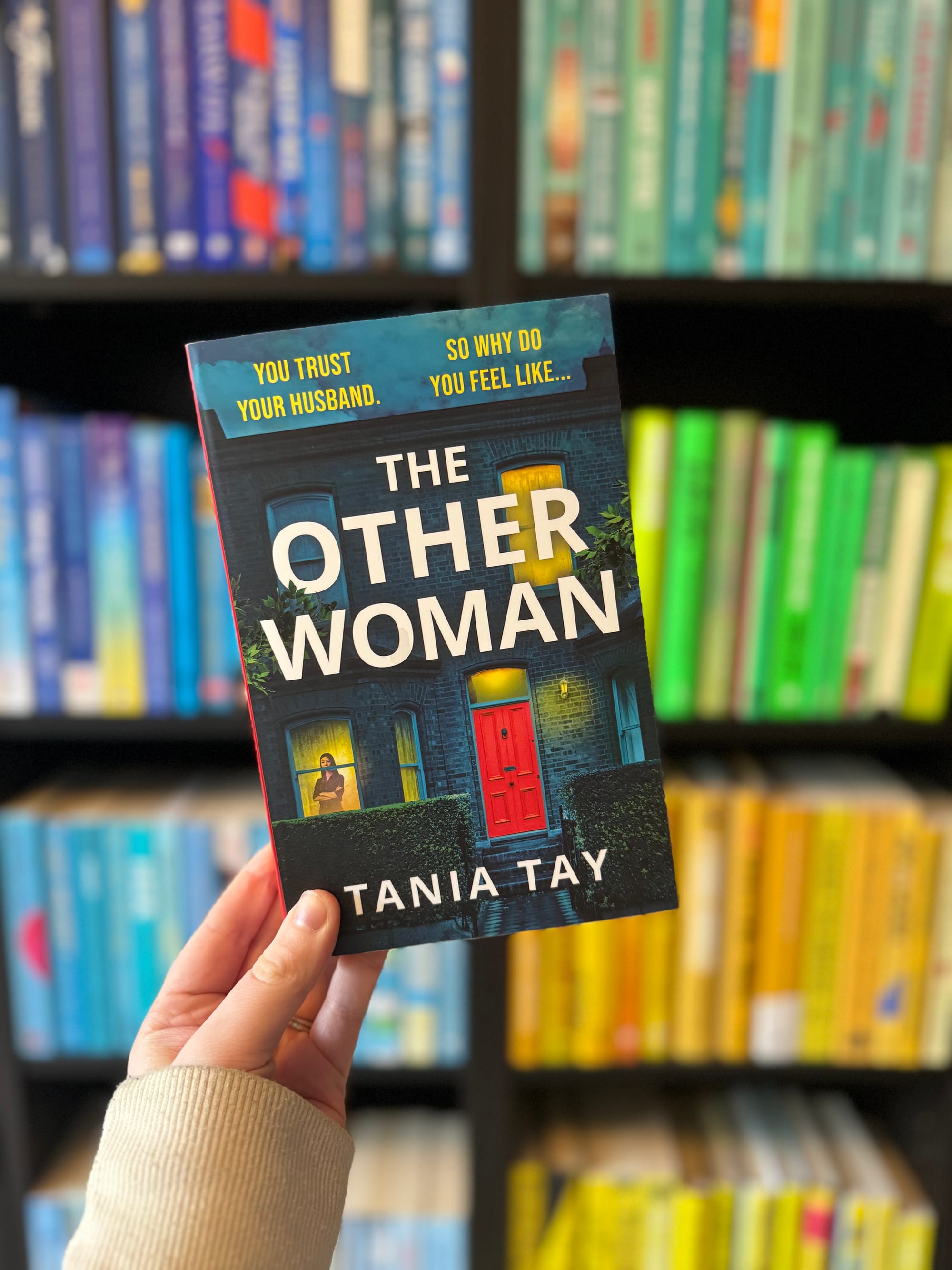 A hand holds "The Other Woman" by Tania Tay. The cover features a red door. Background: colorful books on a shelf. Text: "You trust your husband. So why do you feel like...". Mood: mysterious.
