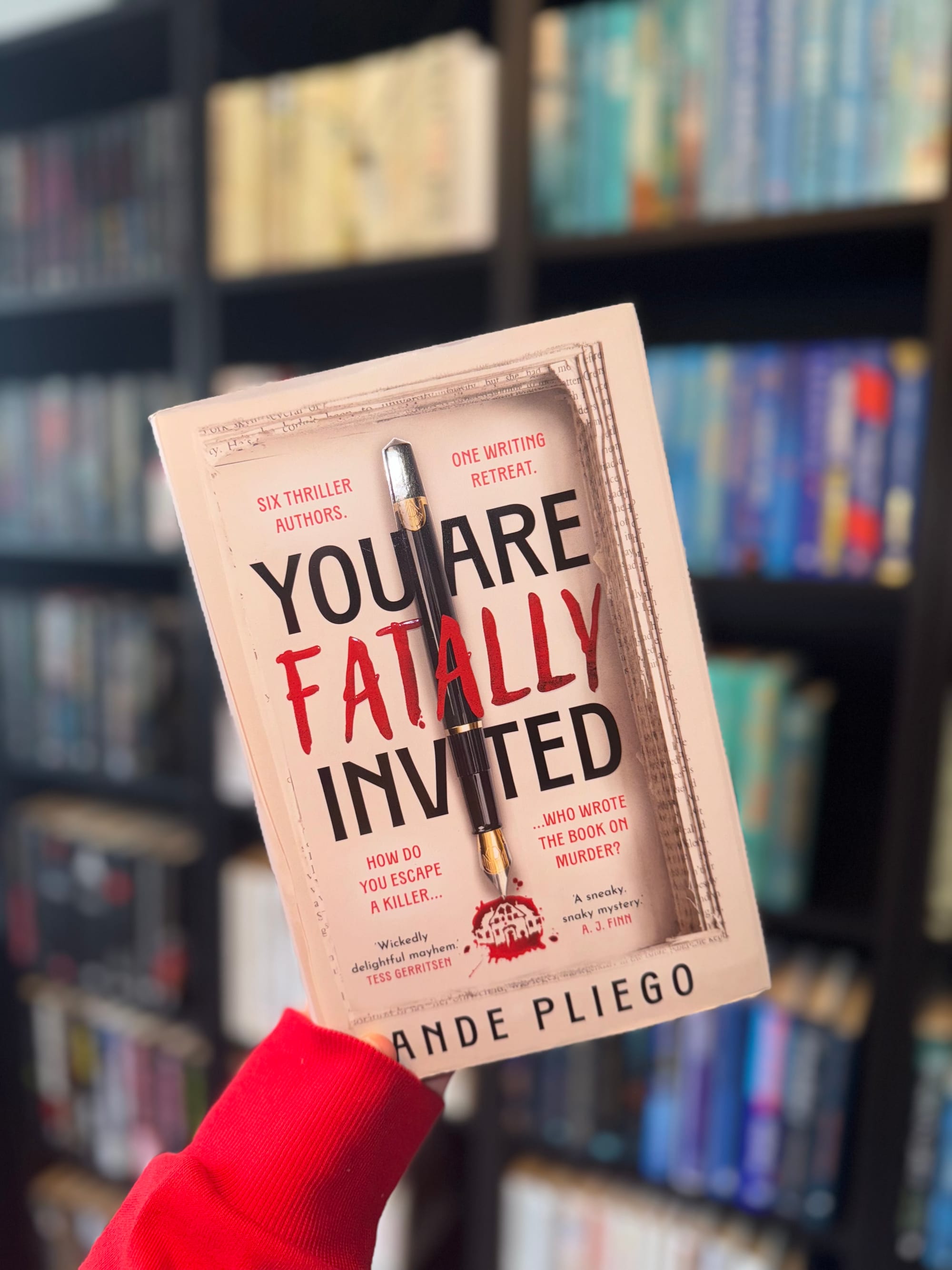 A hand holds a thriller novel, "You Are Fatally Invited," featuring a blood-splattered cover with a pen. Blurred bookshelves are in the background.