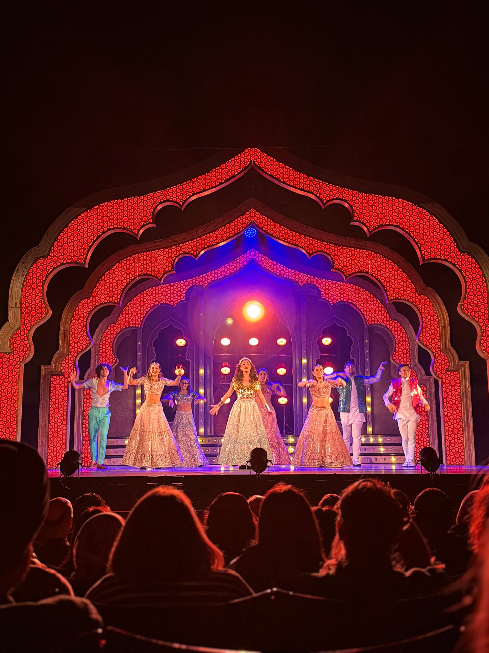 Performers in colorful traditional attire dance on a stage with a vibrant red arch backdrop, illuminated by warm spotlights, before an audience.