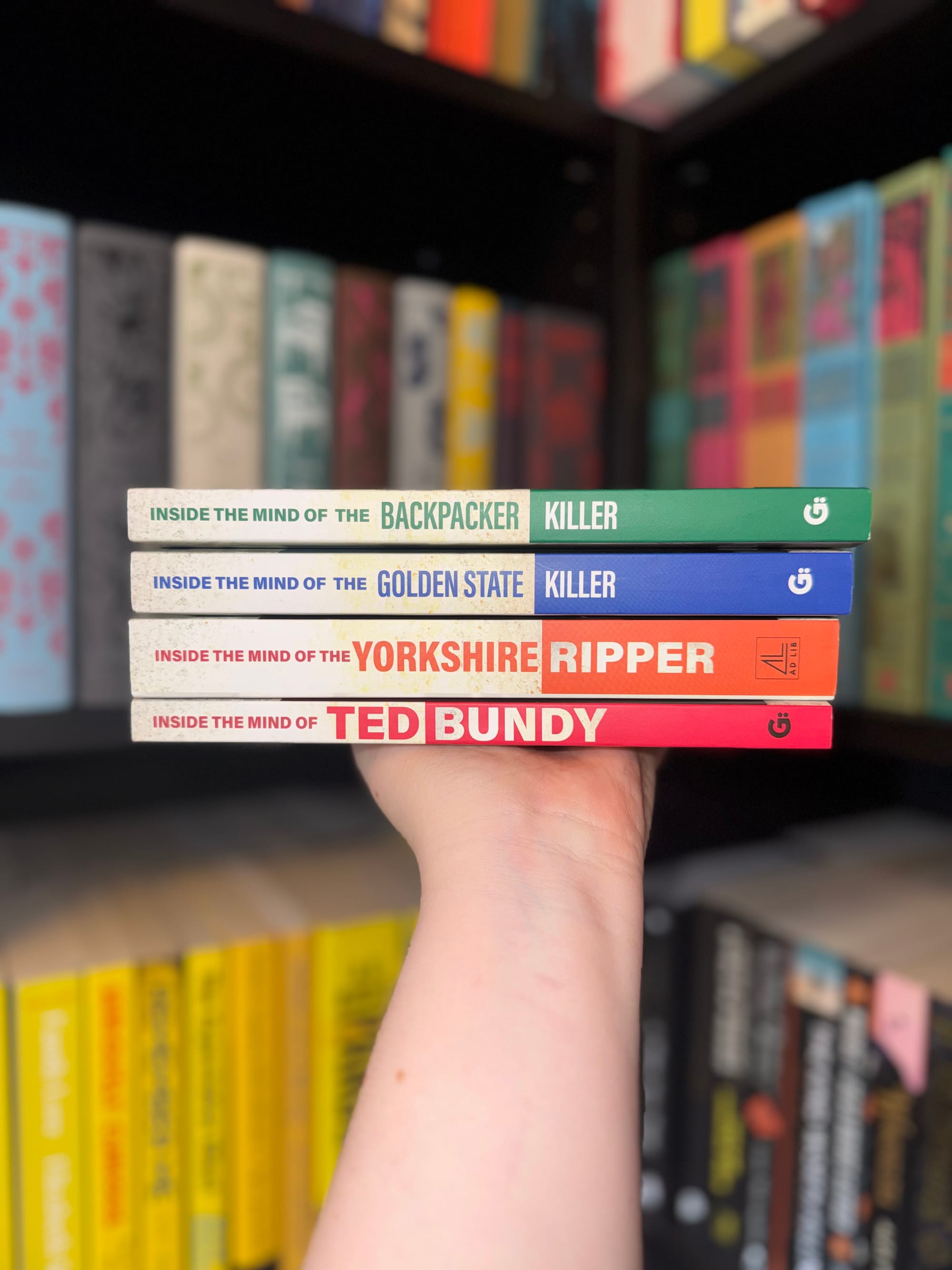 A close-up of four stacked true crime books from the Inside the Mind of the Killer series held in one hand. The spines show the titles: Inside the Mind of the Backpacker Killer, Inside the Mind of the Golden State Killer, Inside the Mind of the Yorkshire Ripper, and Inside the Mind of Ted Bundy, against a backdrop of neatly arranged books on shelves.