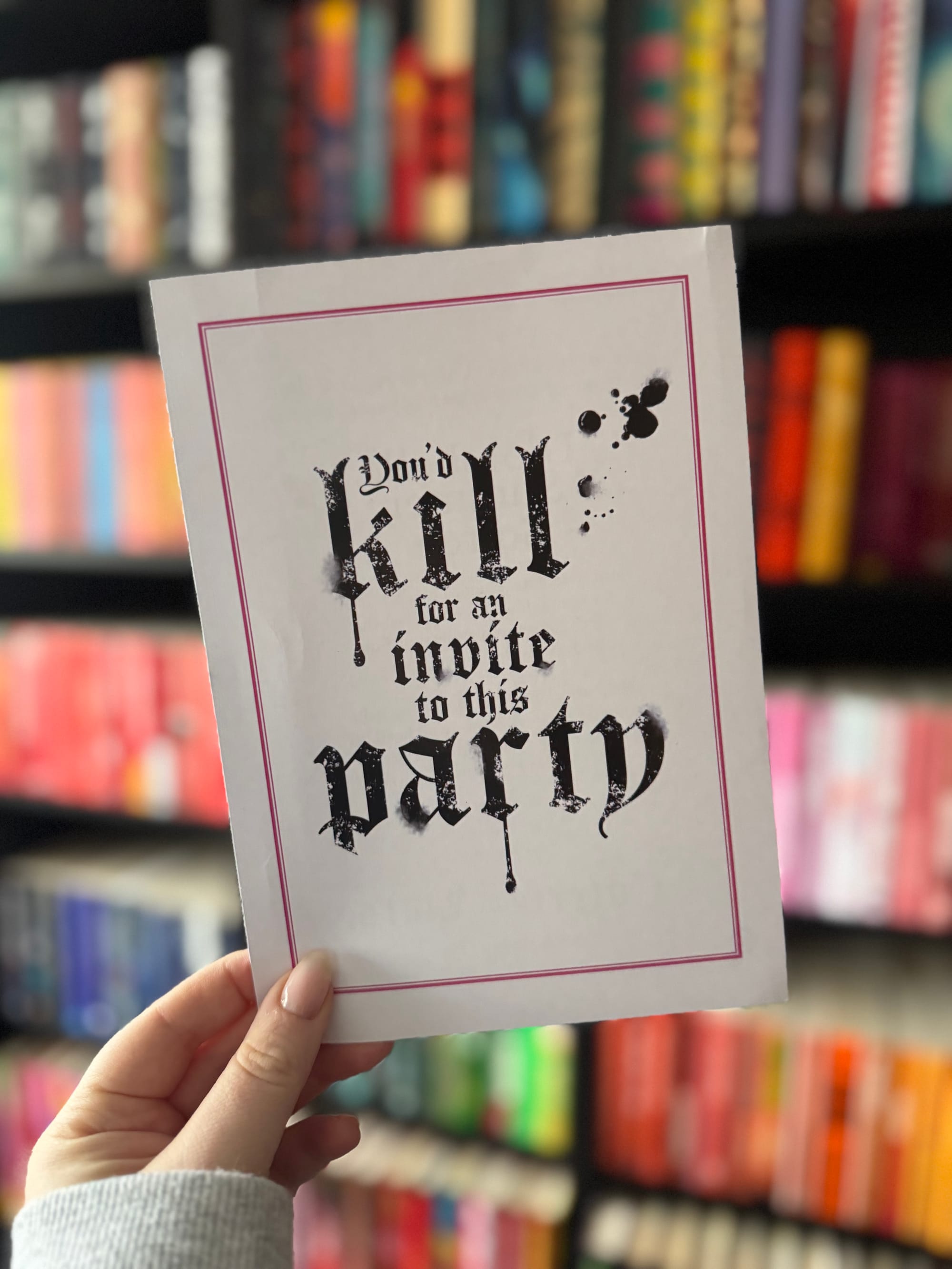 A hand holds a white printed invitation card bordered in pink. In gothic-style black lettering it reads “You’d kill for an invite to this party,” with ink splatter detailing, set against a blurred colourful bookshelf.