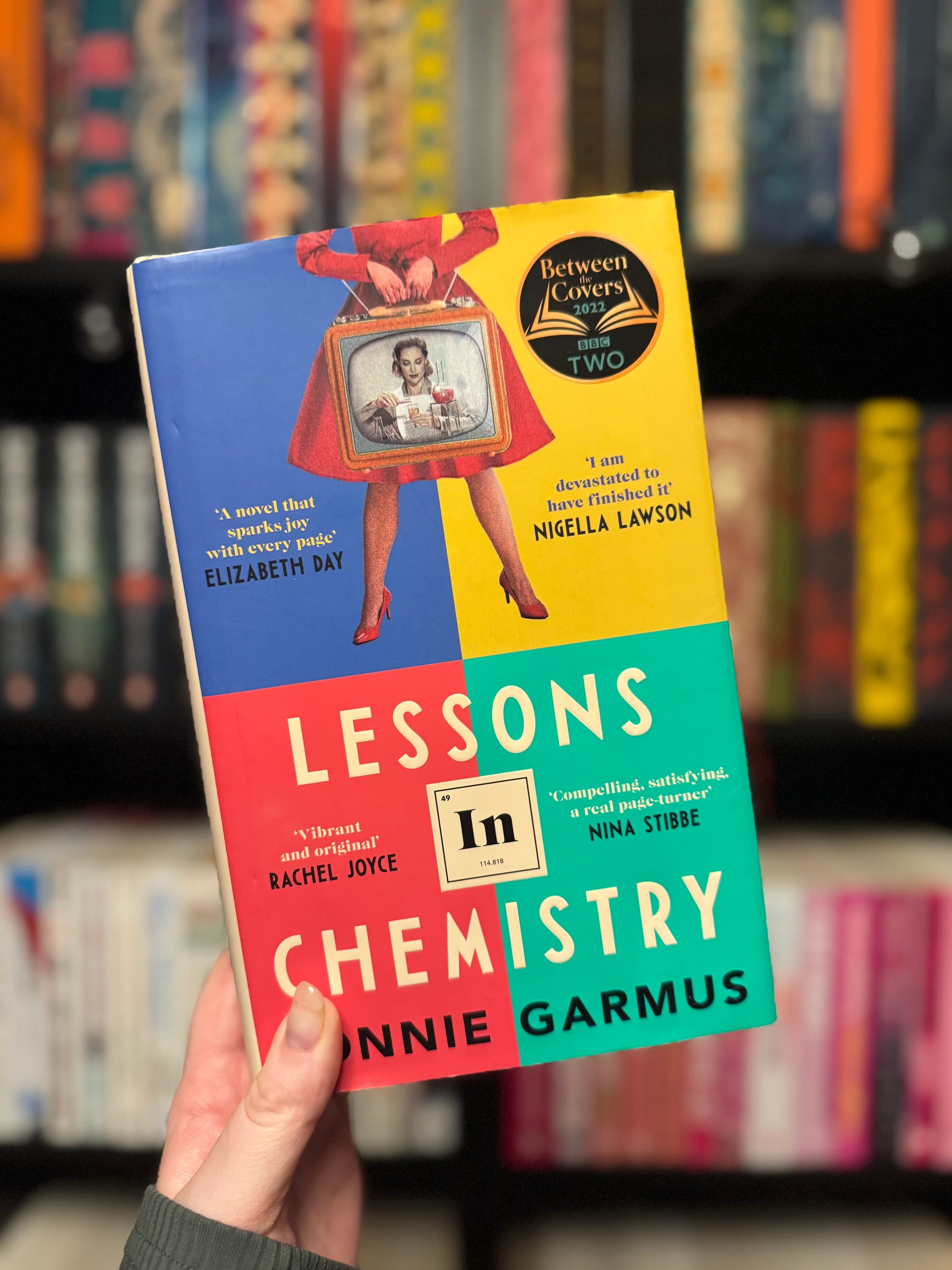 Hand holding the book "Lessons in Chemistry" by Bonnie Garmus against a bookshelf. Cover features a vintage TV illustration and vibrant colors.