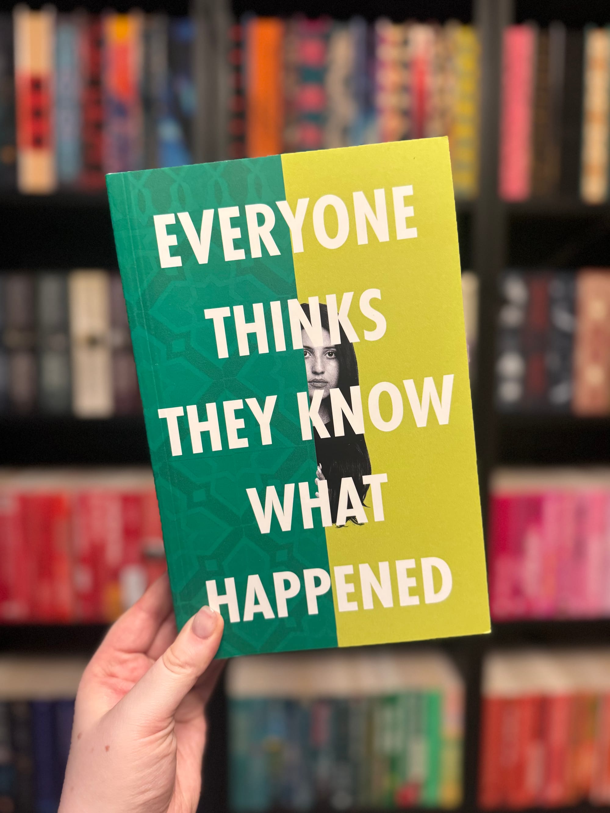 A hand holds a green and yellow book with the title "Everyone Thinks They Know What Happened" against a background of colorful bookshelves.