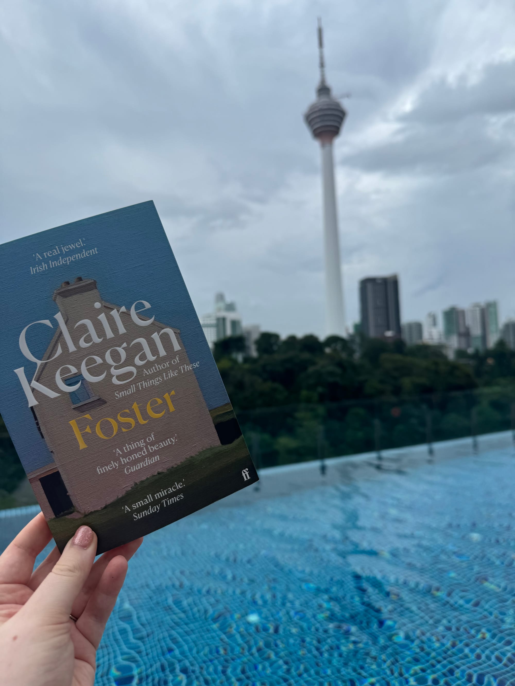Hand holding "Foster" by Claire Keegan above a blue pool, with a cloudy sky and KL tower in the city background.