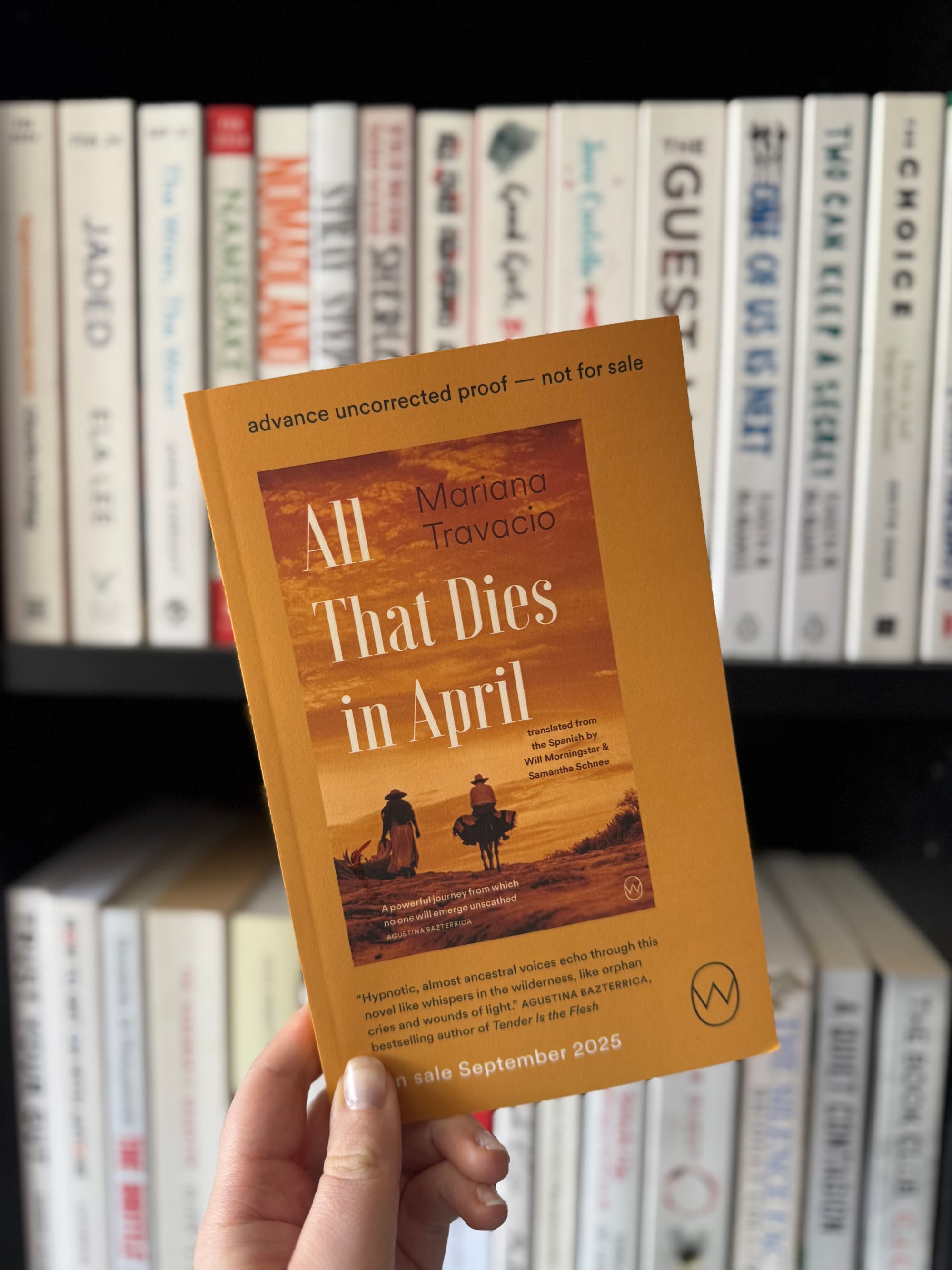 Hand holding a book titled "All That Dies in April" against a blurred bookshelf. The book's cover has a brown tone with figures on horseback.