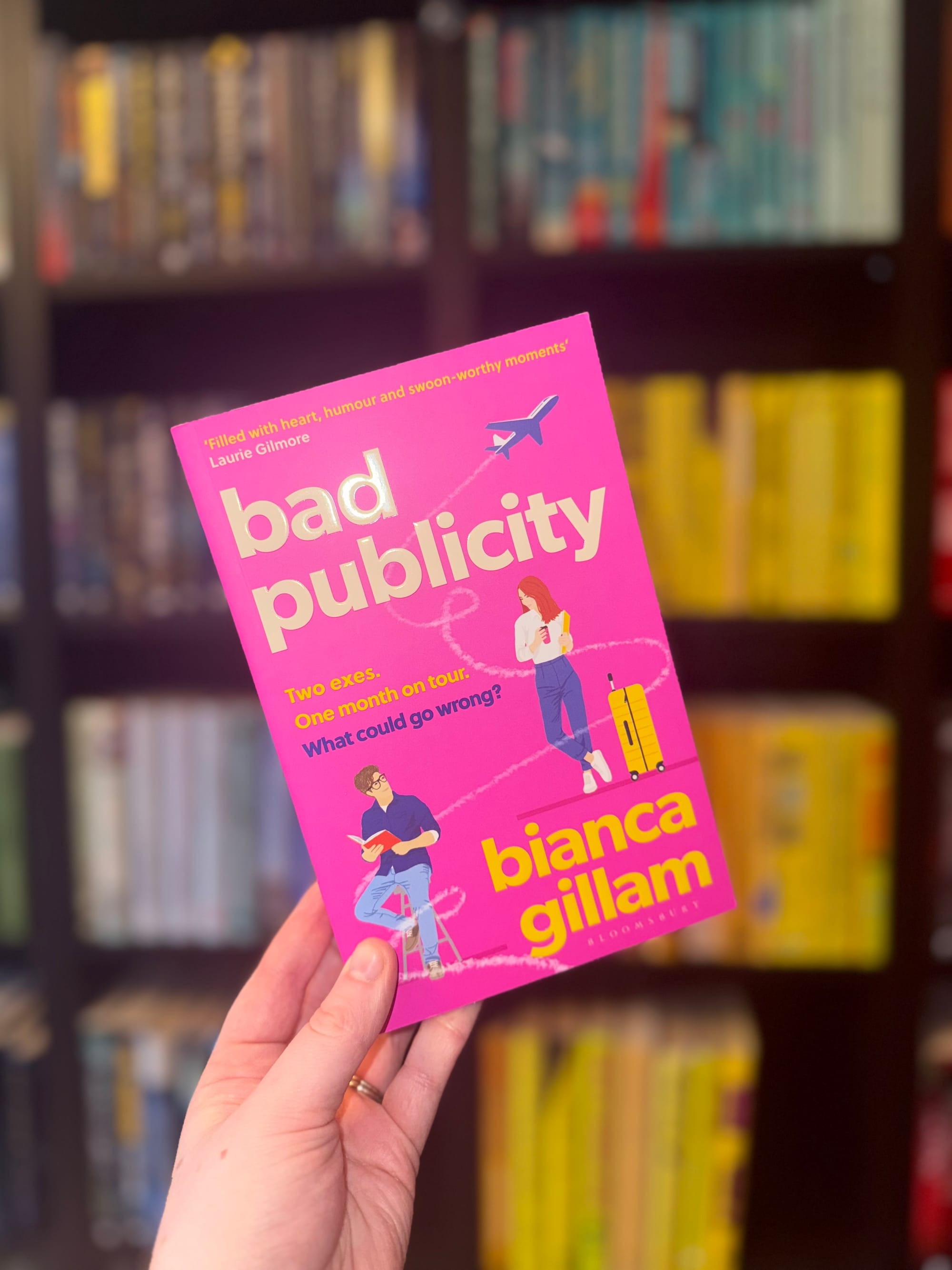 Hand holding "Bad Publicity" book by Bianca Gillam. Bright pink cover with illustrations, airplane graphic, set against a blurred bookshelf.