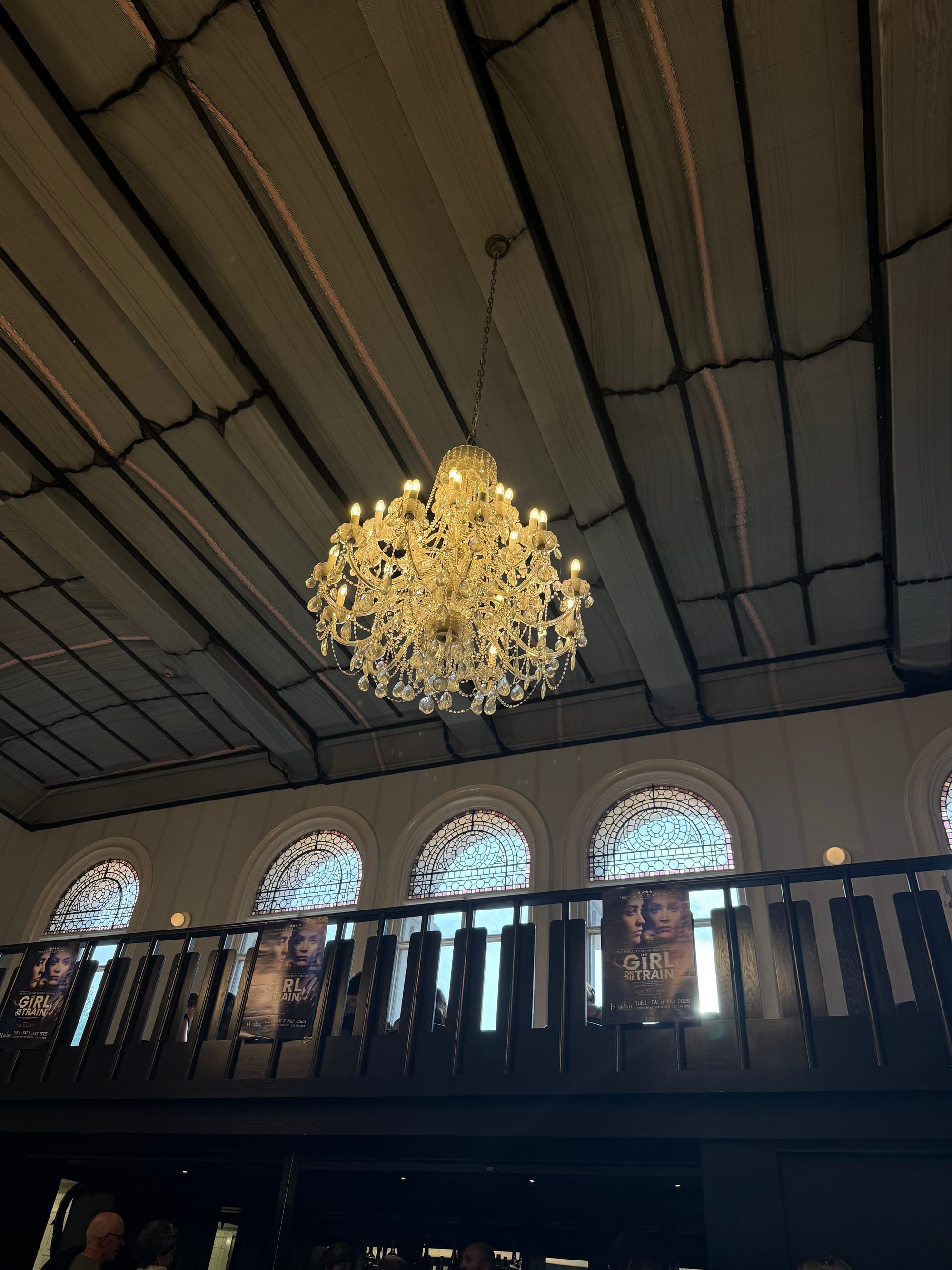 A grand chandelier glows warmly from the high ceiling of Leeds Grand Theatre’s foyer. Below, a balcony displays posters for The Girl on the Train, framed by tall arched windows with stained glass. People gather beneath, enjoying the theatre atmosphere.