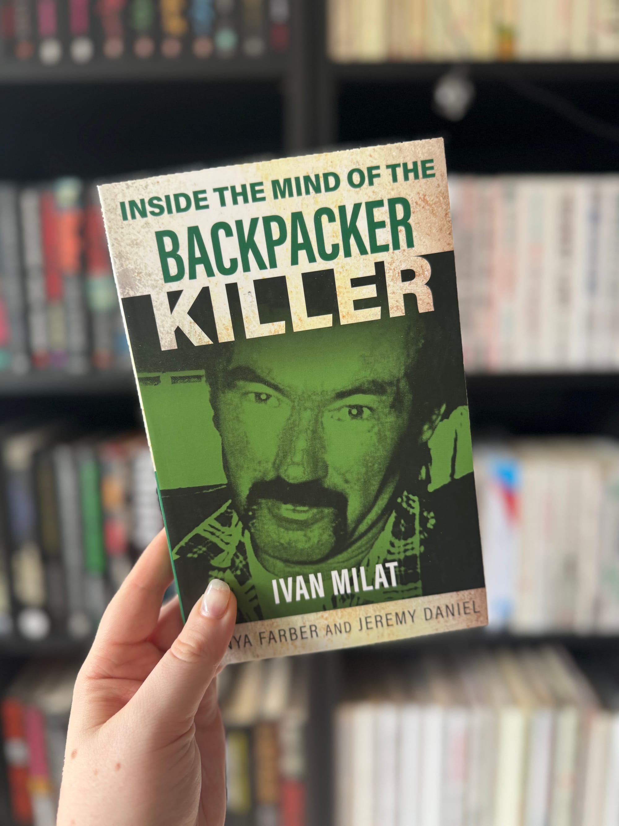 Hand holding a book titled "Inside the Mind of the Backpacker Killer: Ivan Milat" with a green cover. Background shows a blurred bookshelf.