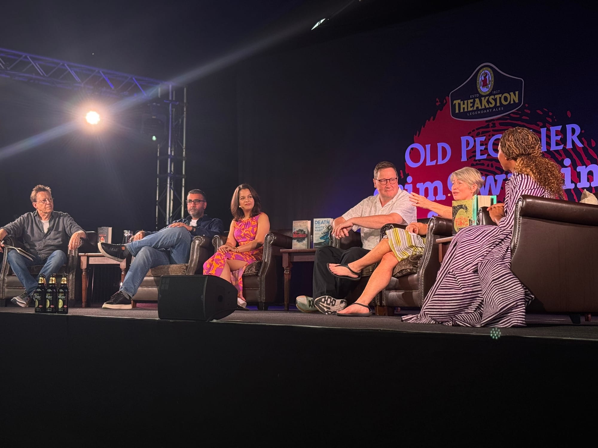 Panel of six people on stage discussing books, with bright spotlight and "Theakston" logo in the background. Participants are relaxed and engaged.