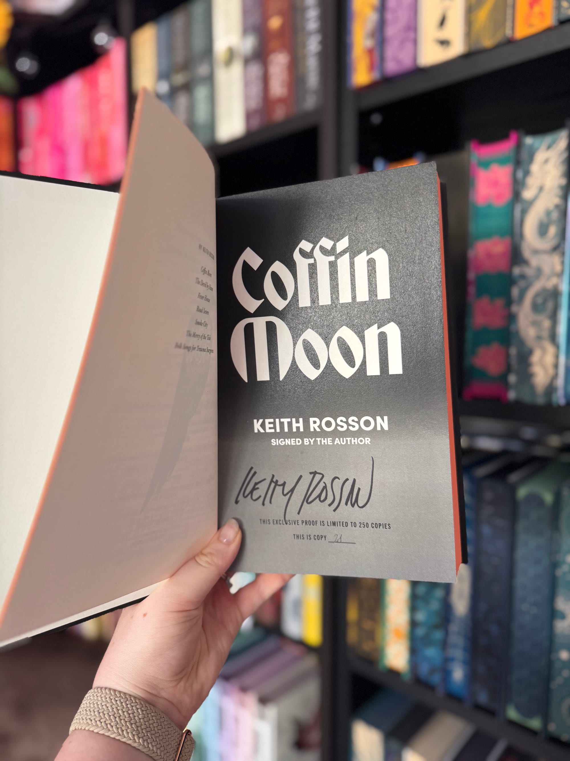 A hand holding open a signed copy of the book 'Coffin Moon' by Keith Rosson. The title page features the author's printed name along with a handwritten signature and a note indicating this is an exclusive proof limited to 250 copies, with this being copy number 34. Colourful bookshelves are visible in the background.