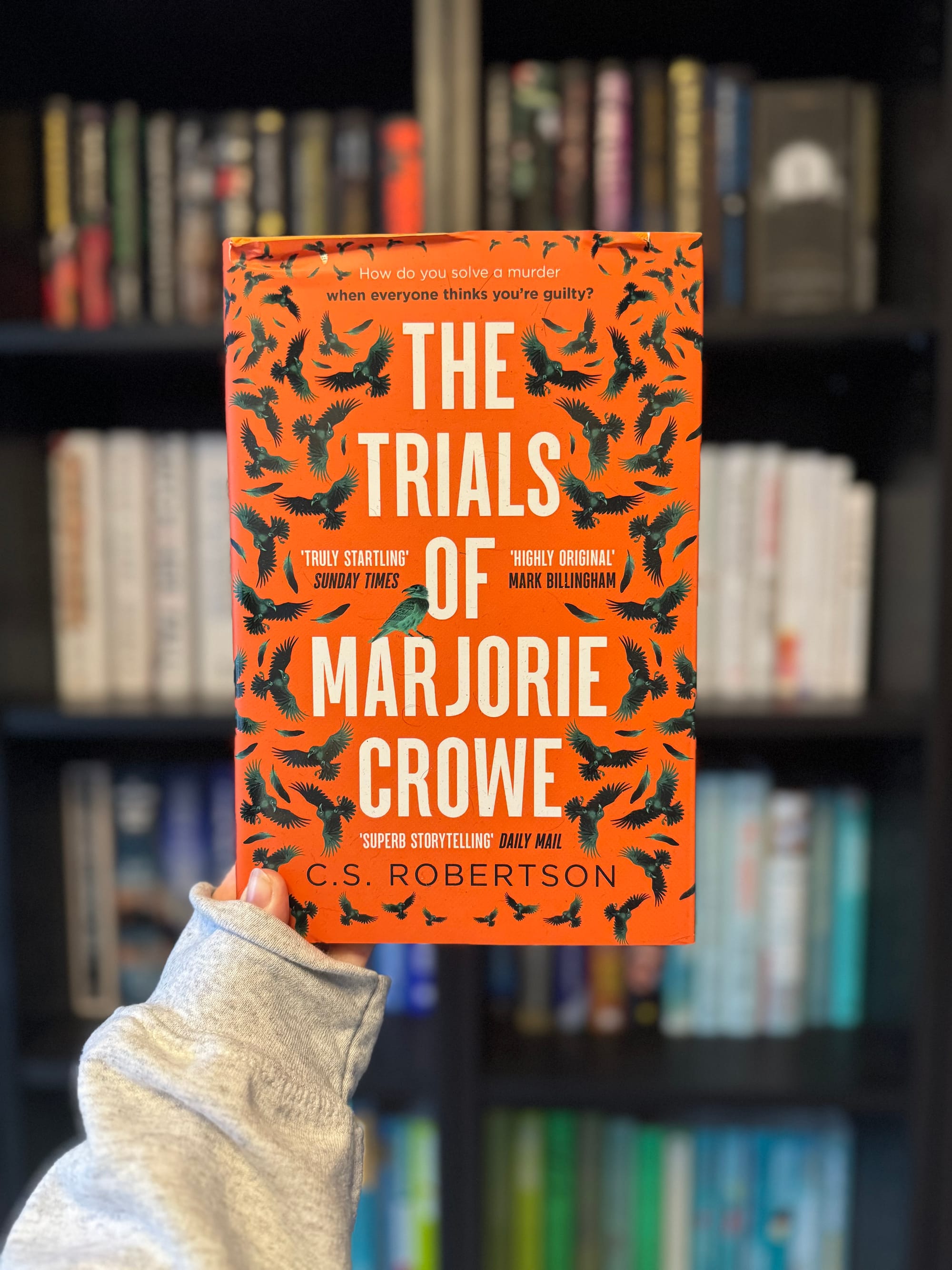 Person holding "The Trials of Marjorie Crowe" book with orange cover and bird patterns. Background shows a blurred bookshelf.