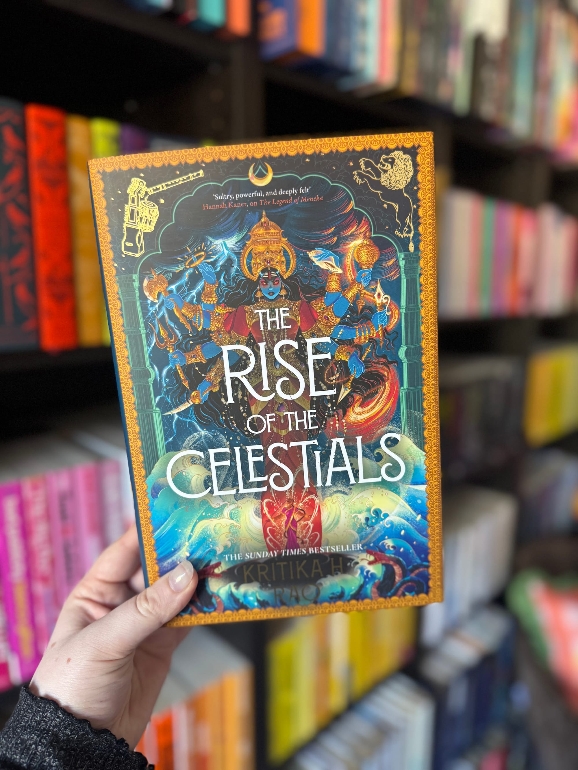 A hand holds "The Rise of the Celestials" book with vibrant cover art in a bookstore. Shelves of colorful books are blurred in the background.