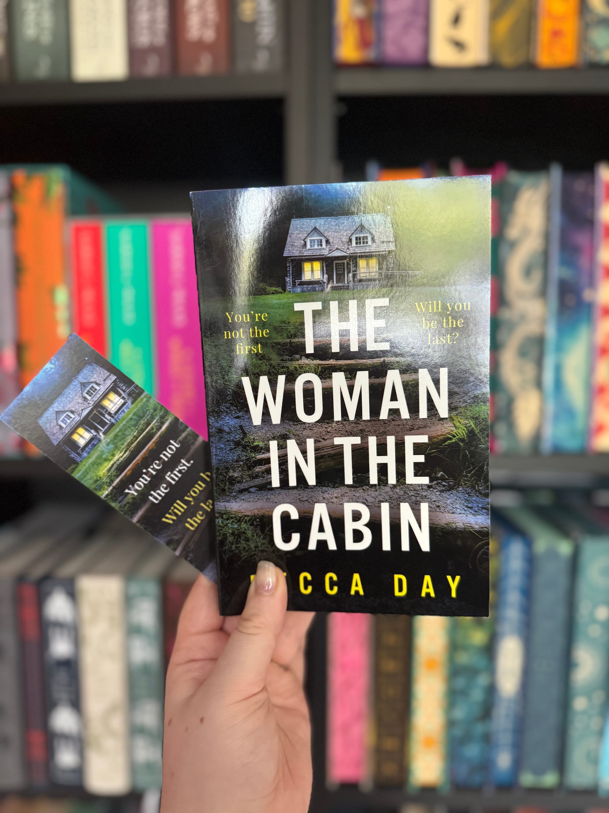 A hand holding the book The Woman in the Cabin by Rebecca Day in front of a colourful bookshelf. The book cover shows a cabin surrounded by greenery under a cloudy sky, with the tagline “You’re not the first. Will you be the last?” A matching bookmark with the same design is also being held alongside the book.