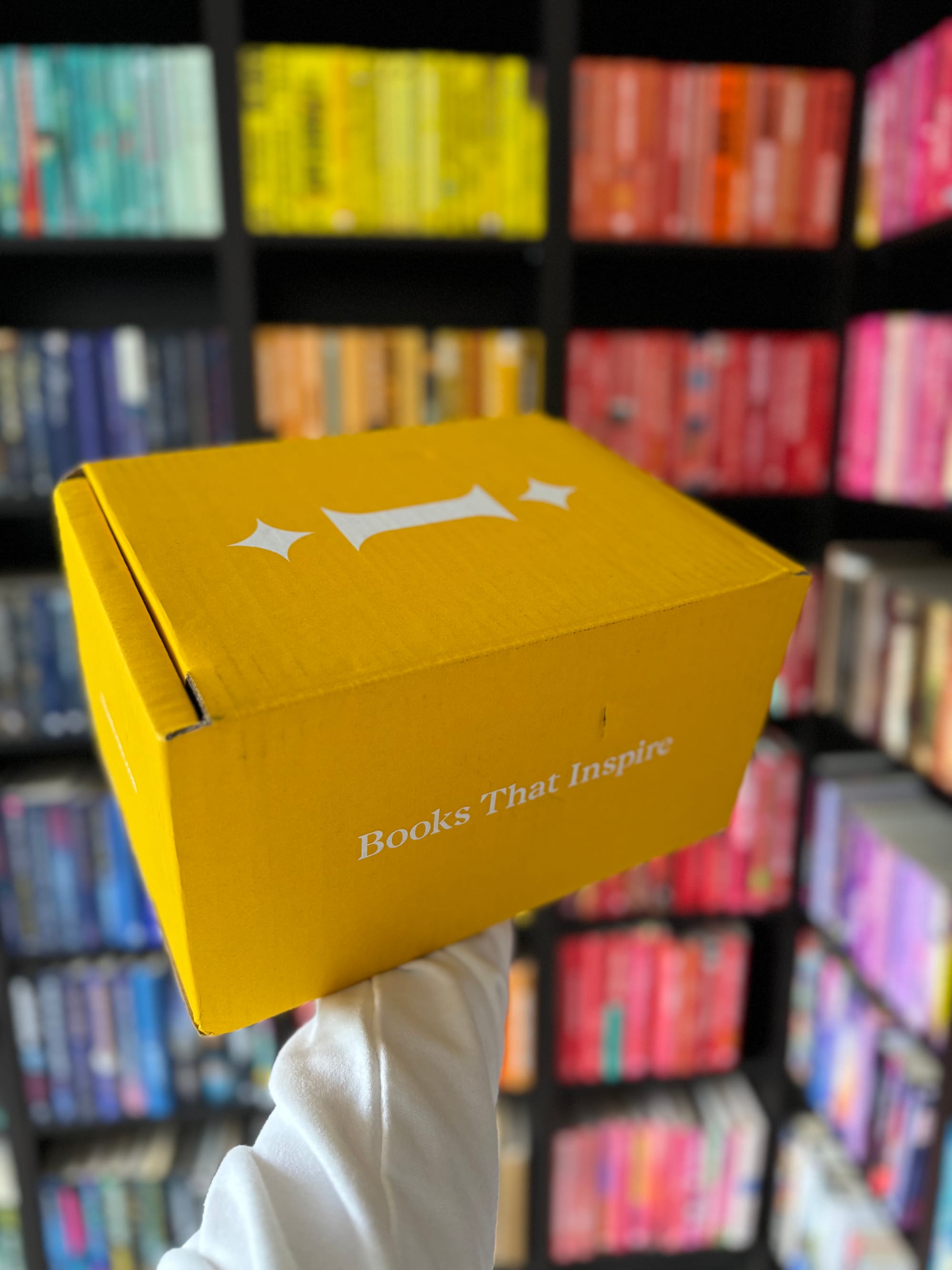 A hand holds a yellow box labeled "Books That Inspire" against shelves of colorful, arranged books, creating a vibrant, organized backdrop.