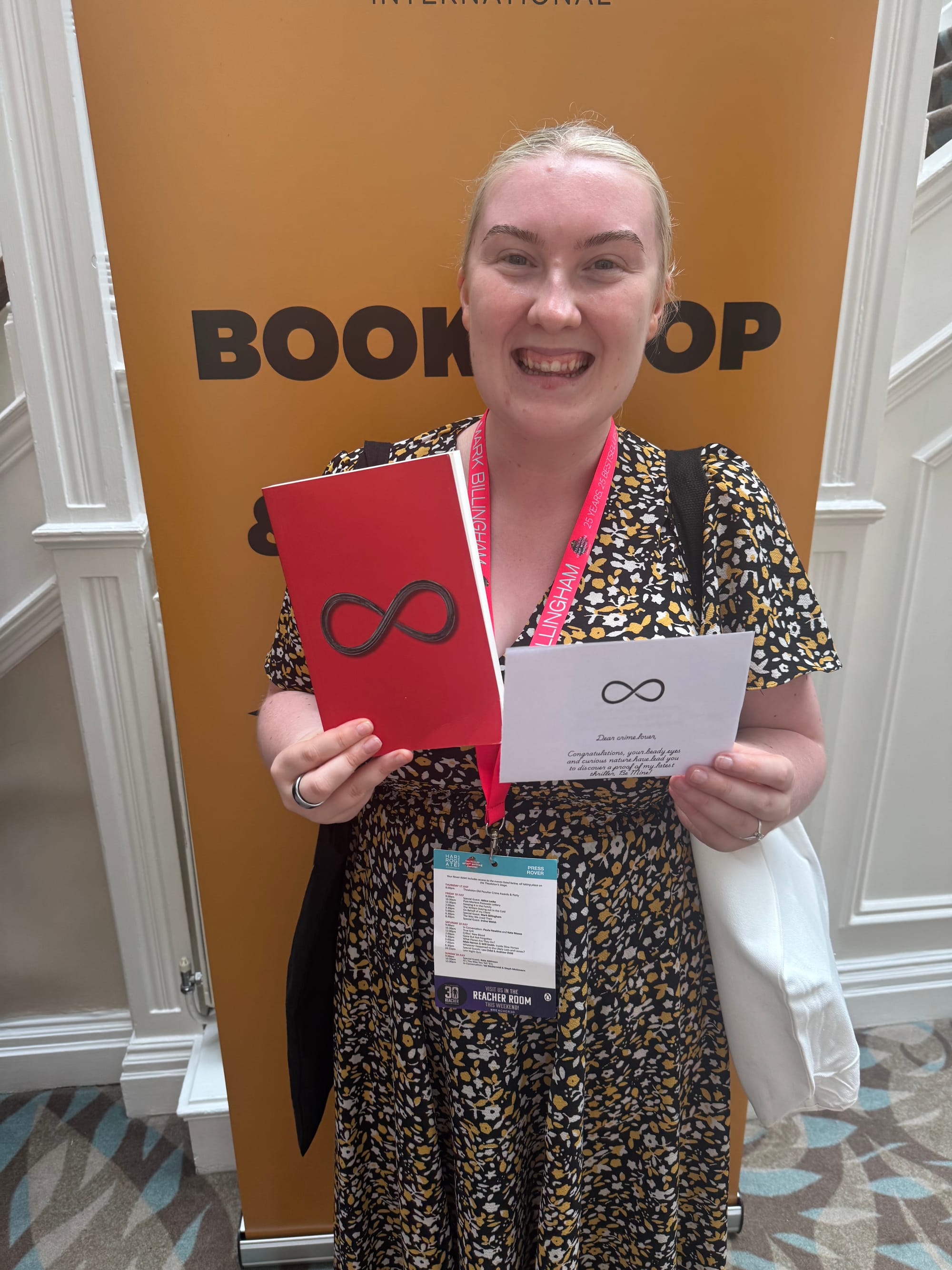 Smiling woman standing indoors holding a red book and envelope from a giveaway, wearing a floral dress and festival lanyard, in front of a “Bookshop” branded backdrop.