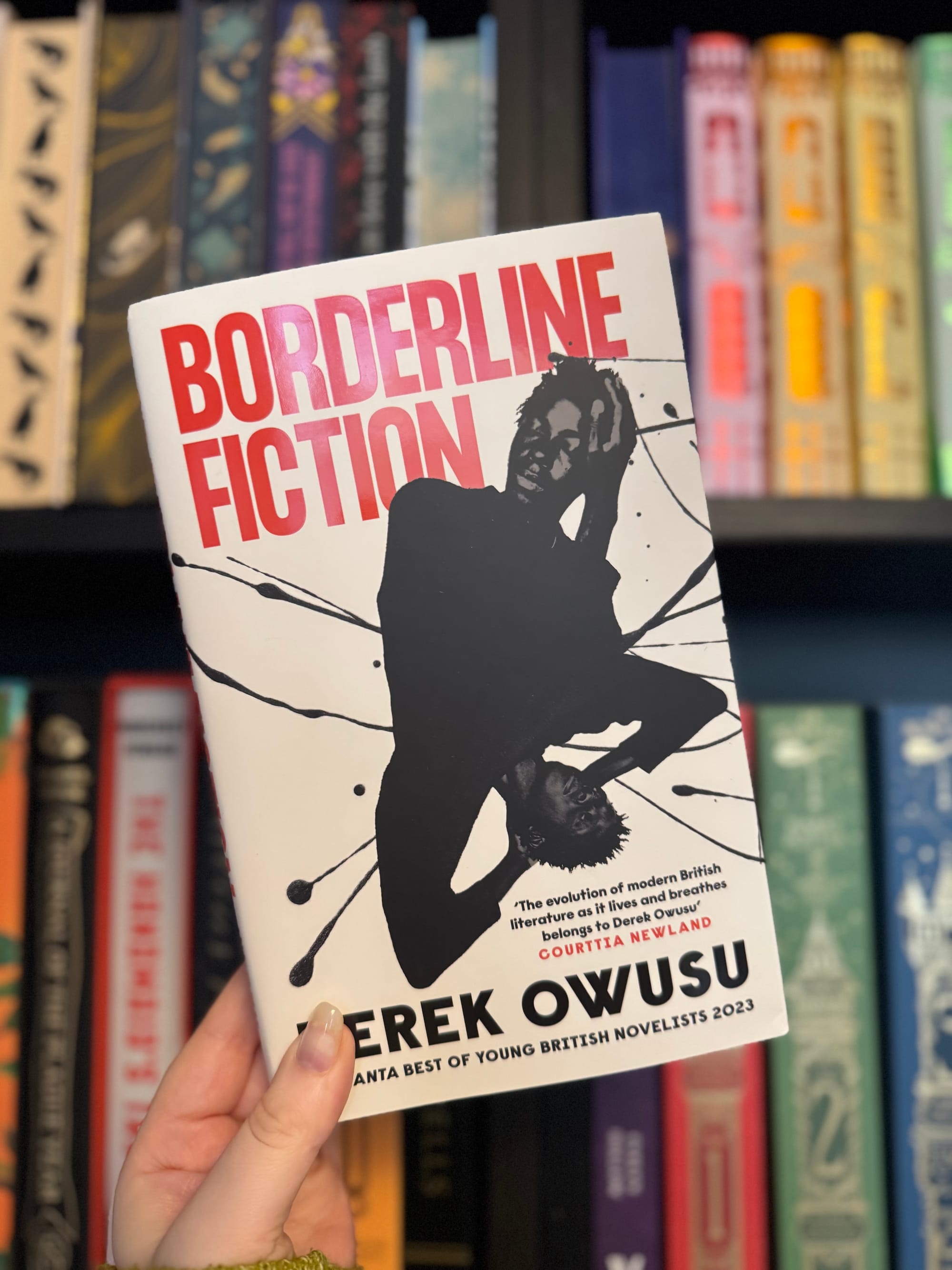 Hand holds "Borderline Fiction" book by Derek Owusu. Bold red and black design with a silhouette. Bookshelf background with colorful spines.