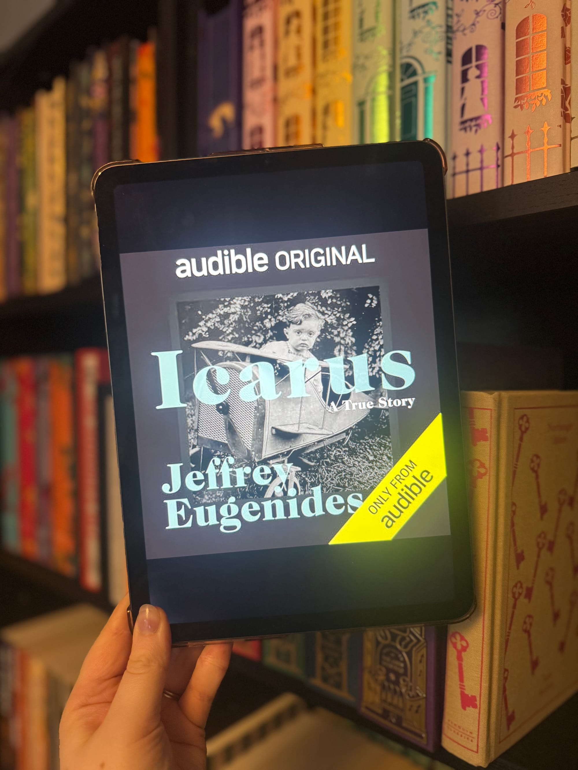 Hand holds a tablet displaying an Audible cover for "Icarus" by Jeffrey Eugenides. Background shows colorful, patterned bookshelves.