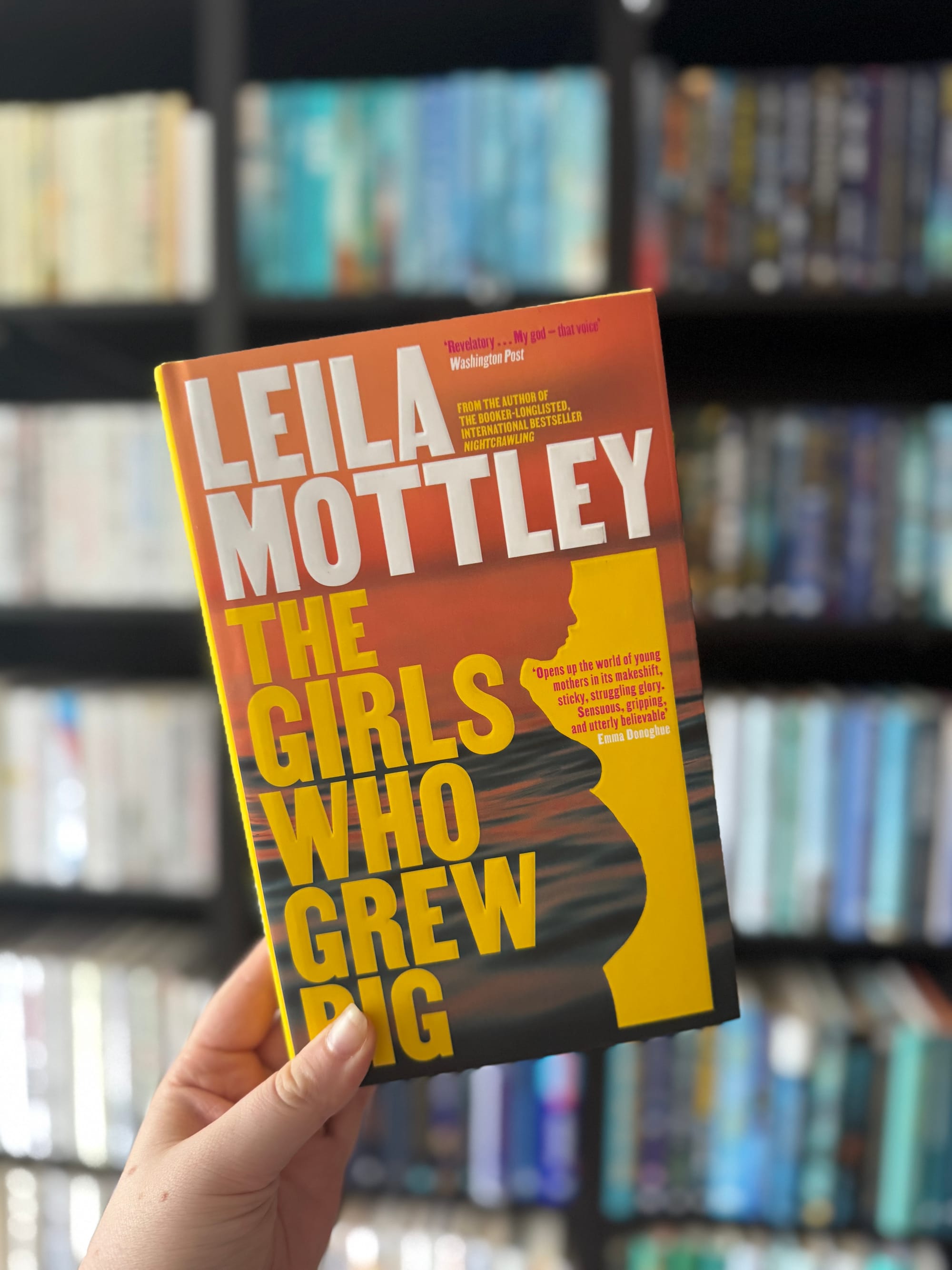Hand holds "The Girls Who Grew Big" by Leila Mottley, with a silhouette cover. Bookshelf with blurred books in the background.