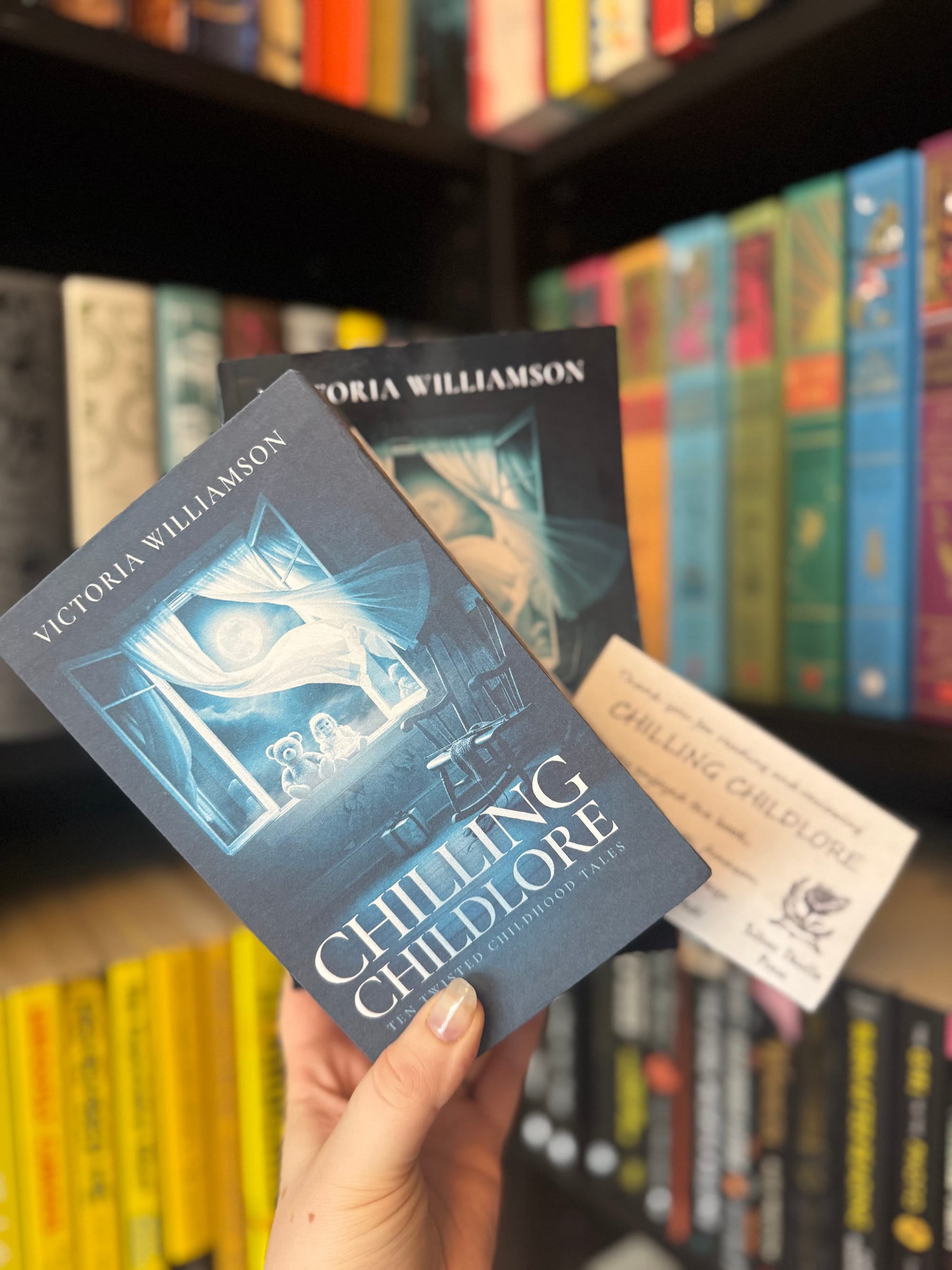 A hand holds the book Chilling Childlore along with a Silver Thistle Press card, in front of a bookshelf with colourful spines. The eerie blue cover art featuring moonlight and a shadowy room is clearly visible.
