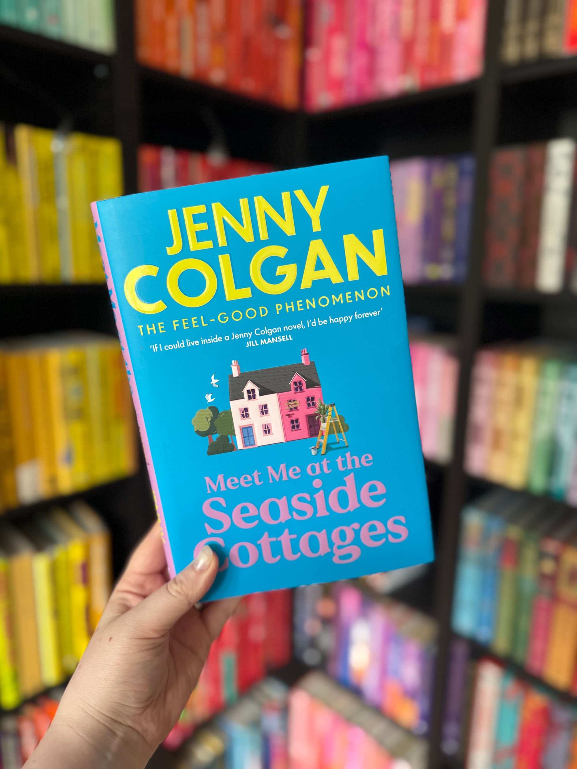 A hand holds the book "Meet Me at the Seaside Cottage" by Jenny Colgan. Blue cover with pink cottages. Colorful bookshelves in the background.