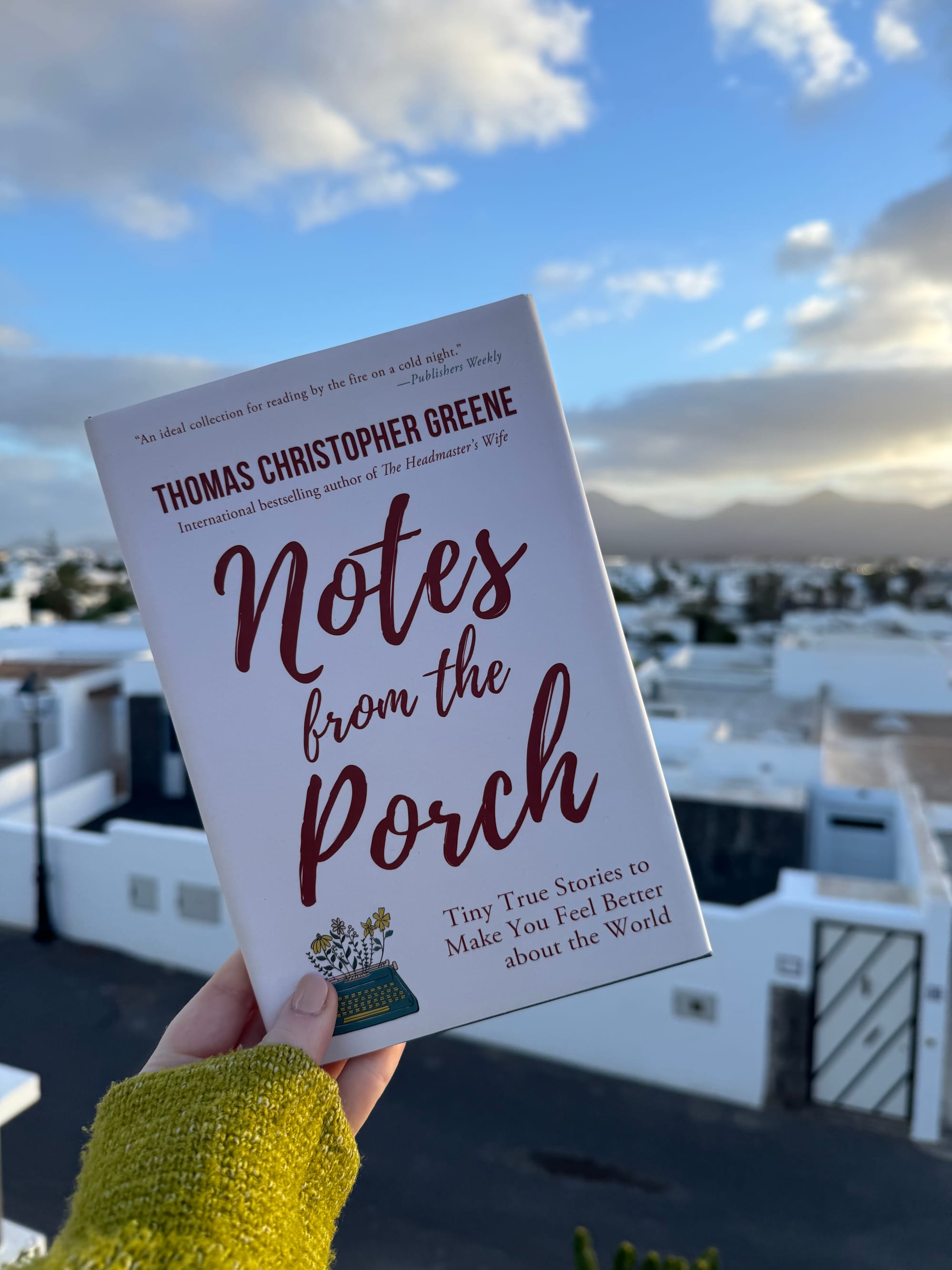 Hand holding a book titled "Notes from the Porch" against a backdrop of a cloudy blue sky and white buildings, creating a tranquil mood.