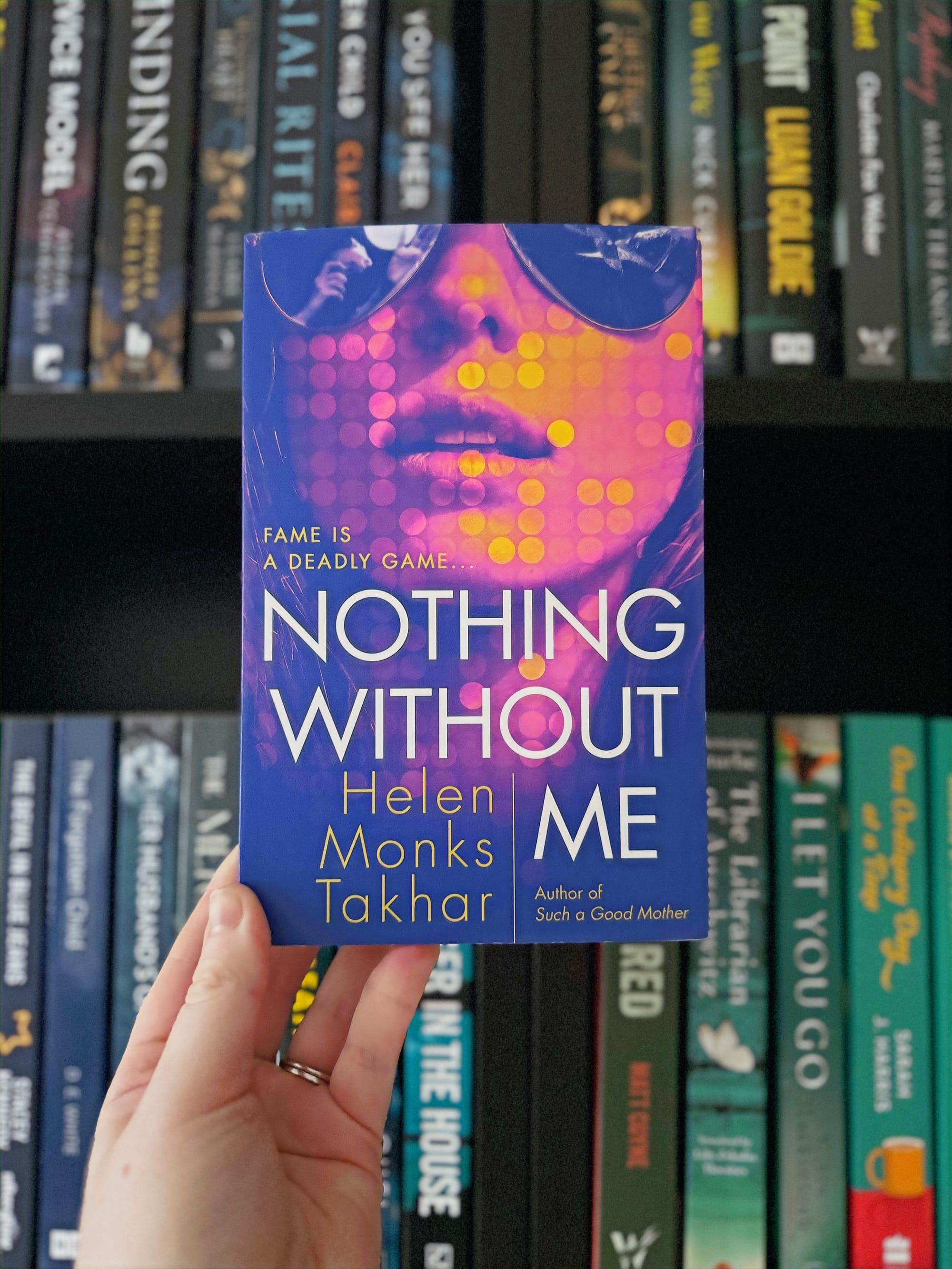 Hand holding the book "Nothing Without Me" by Helen Monks Takhar, set against a bookshelf background. The cover is vibrant with pink and yellow dots.