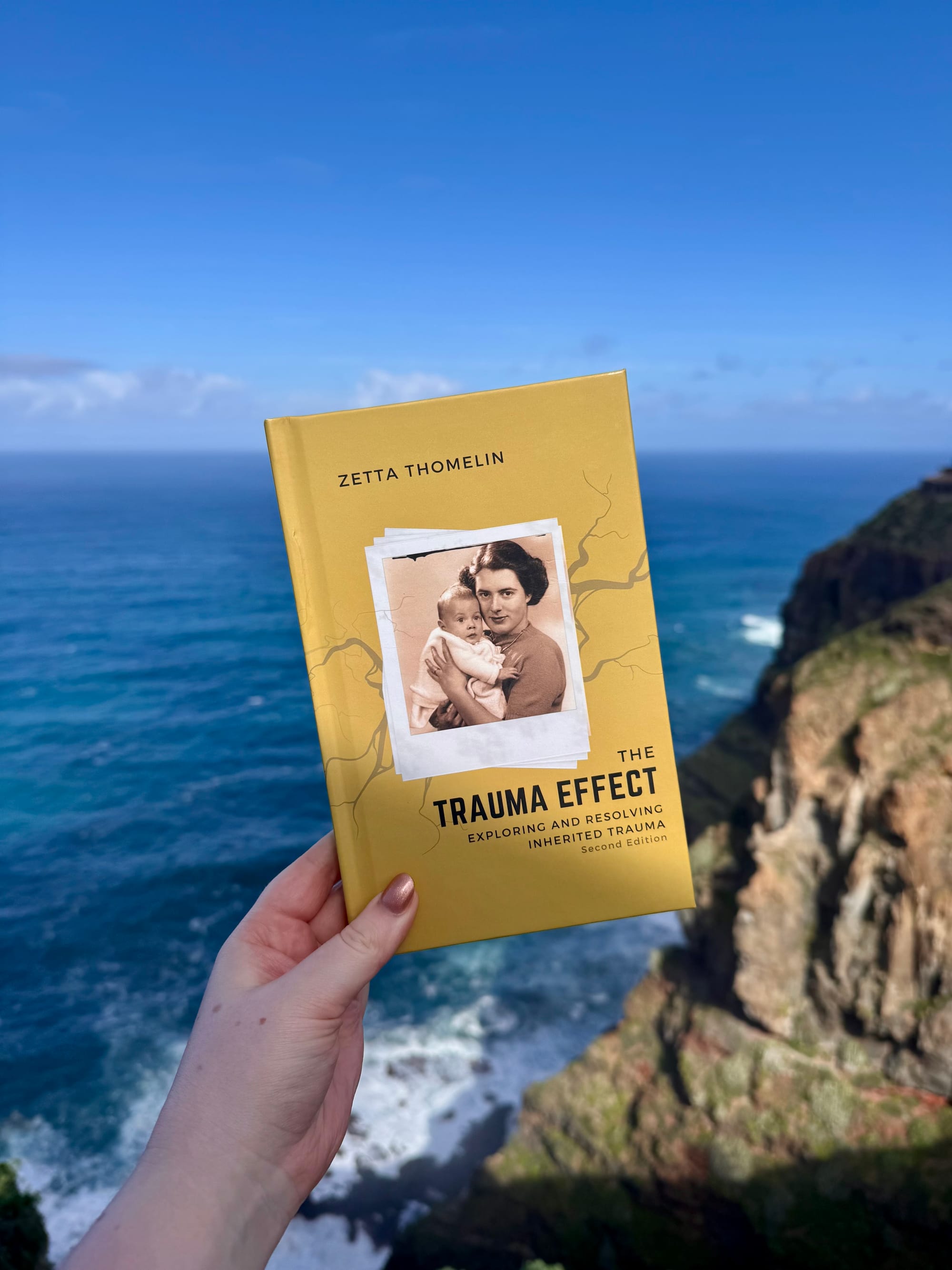 Hand holds yellow book "The Trauma Effect" with a photo on the cover. Background shows a scenic ocean view and cliffs under a clear blue sky.