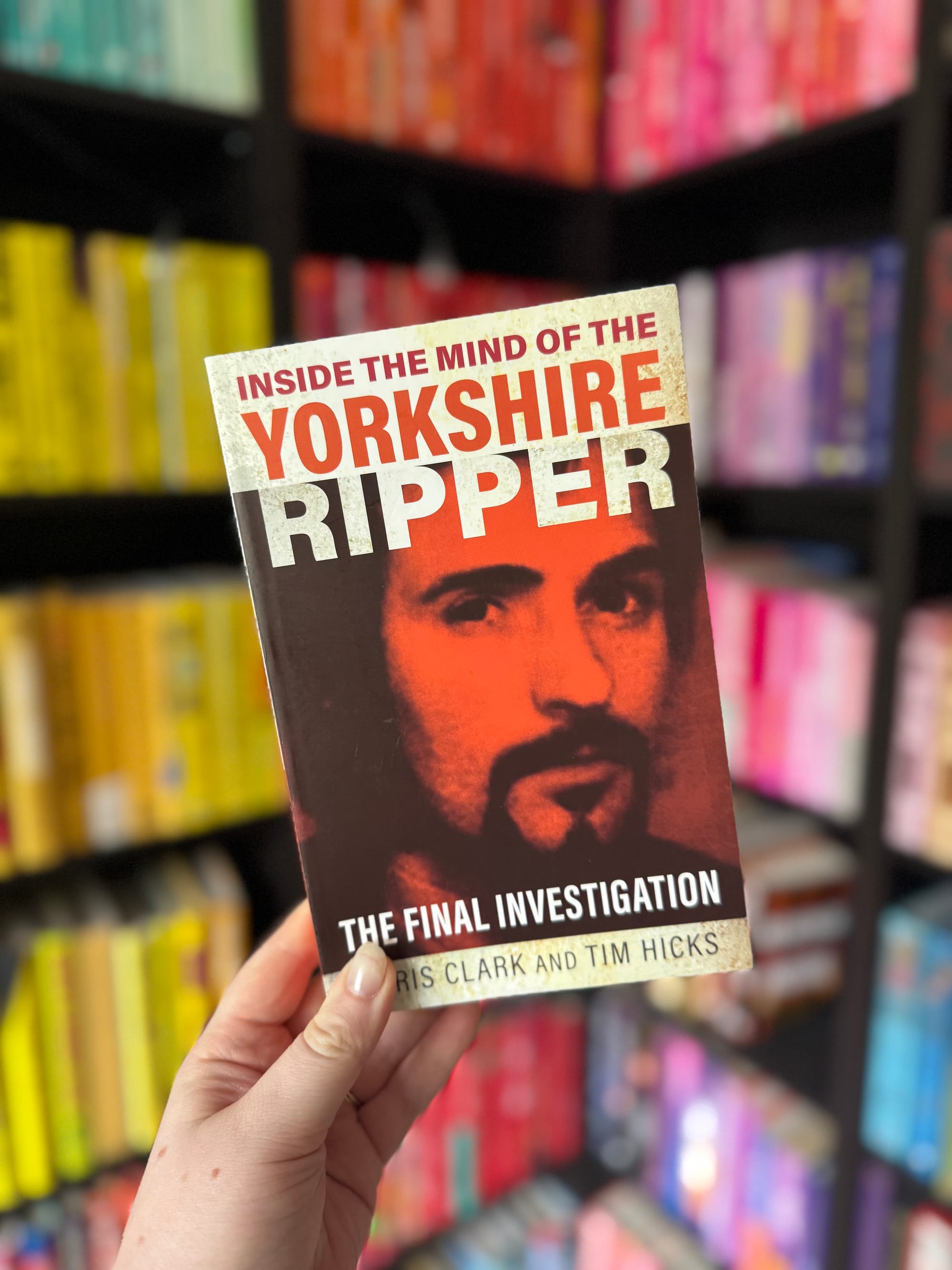 Hand holding a book titled "Inside the Mind of the Yorkshire Ripper" in a colorful bookstore with shelves of various hues in the background.