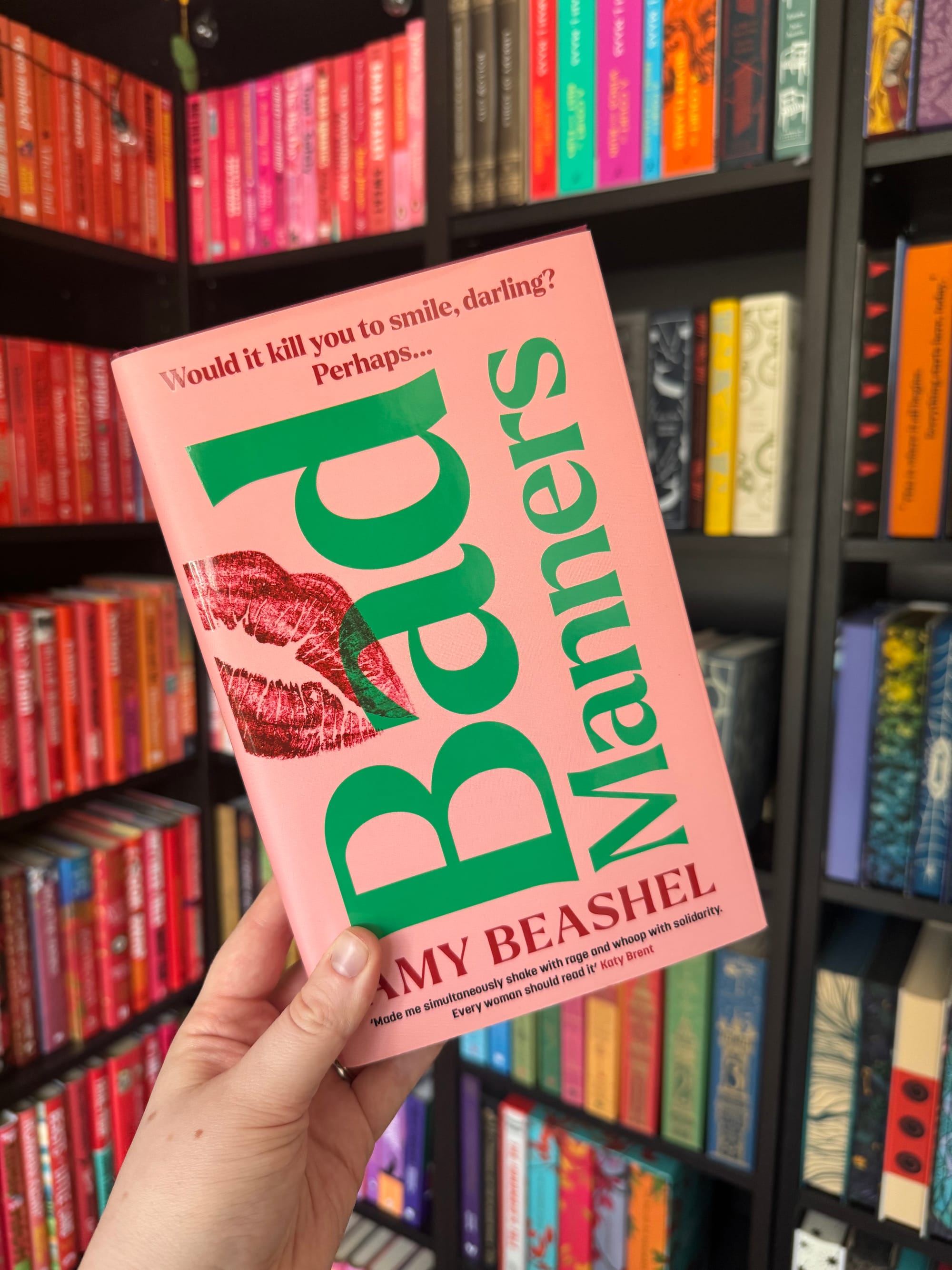 Hand holding a pink book titled "Bad Manners" by Amy Beashel in a colorful library. Red lipstick print and playful text on the cover.