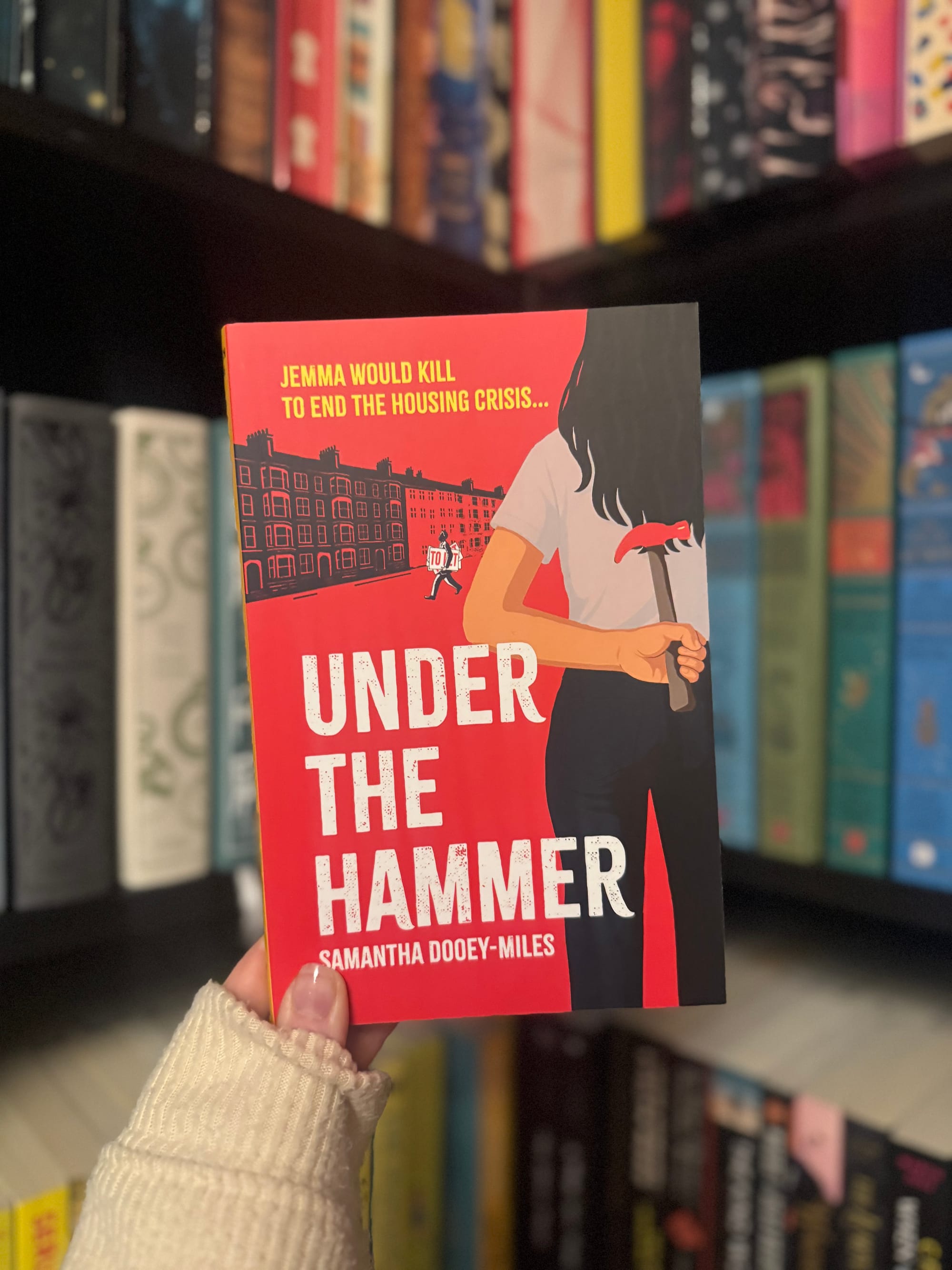 A hand holds a red book titled "Under the Hammer" by Samantha Dooey-Miles. The background shows colorful books on a bookshelf.