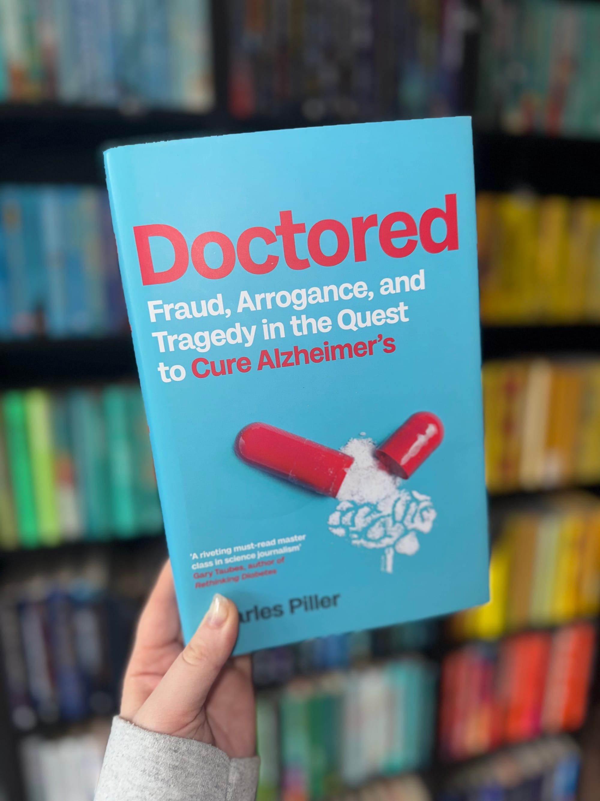 Hand holding the book "Doctored" with a blue cover, red pill graphic, and text about Alzheimer's. Background: blurred bookshelves.