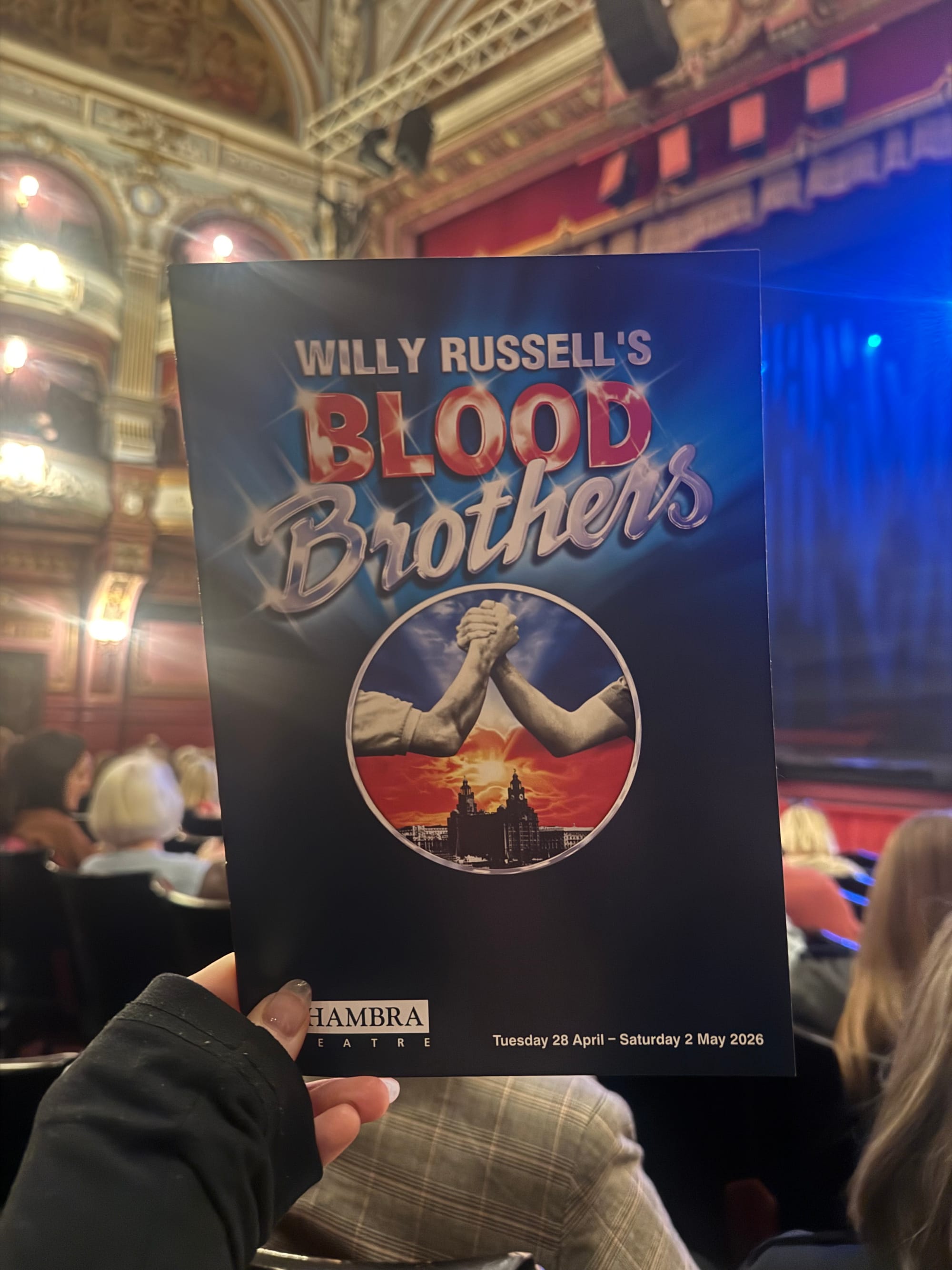 Hand holding a "Blood Brothers" program in a theater. Ornate interior with red and gold accents visible. Show dates are April 28-May 2, 2026.