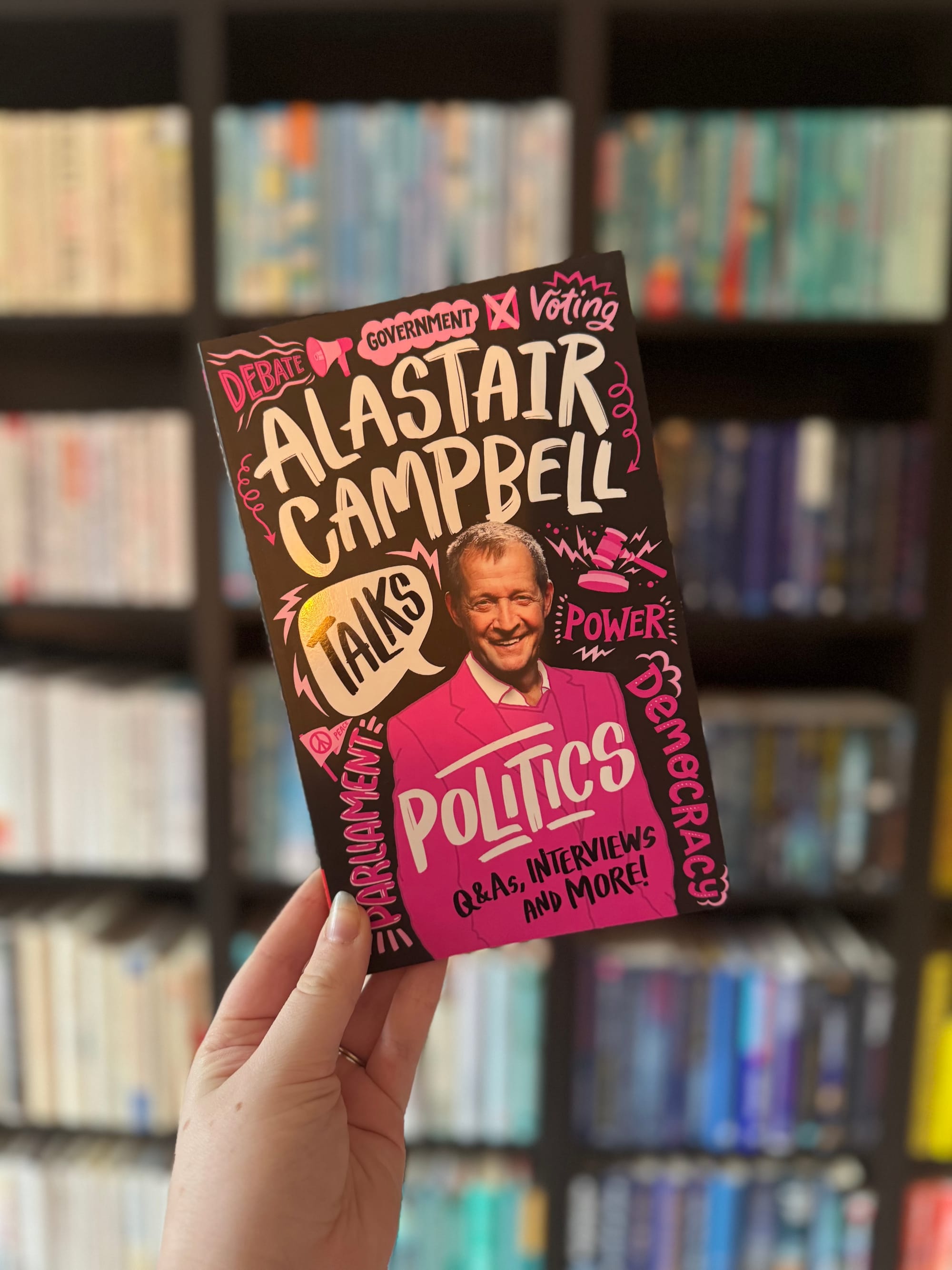 Hand holding a colorful book titled "Alastair Campbell Talks Politics" against a blurred bookshelf background. Text includes debates, interviews.