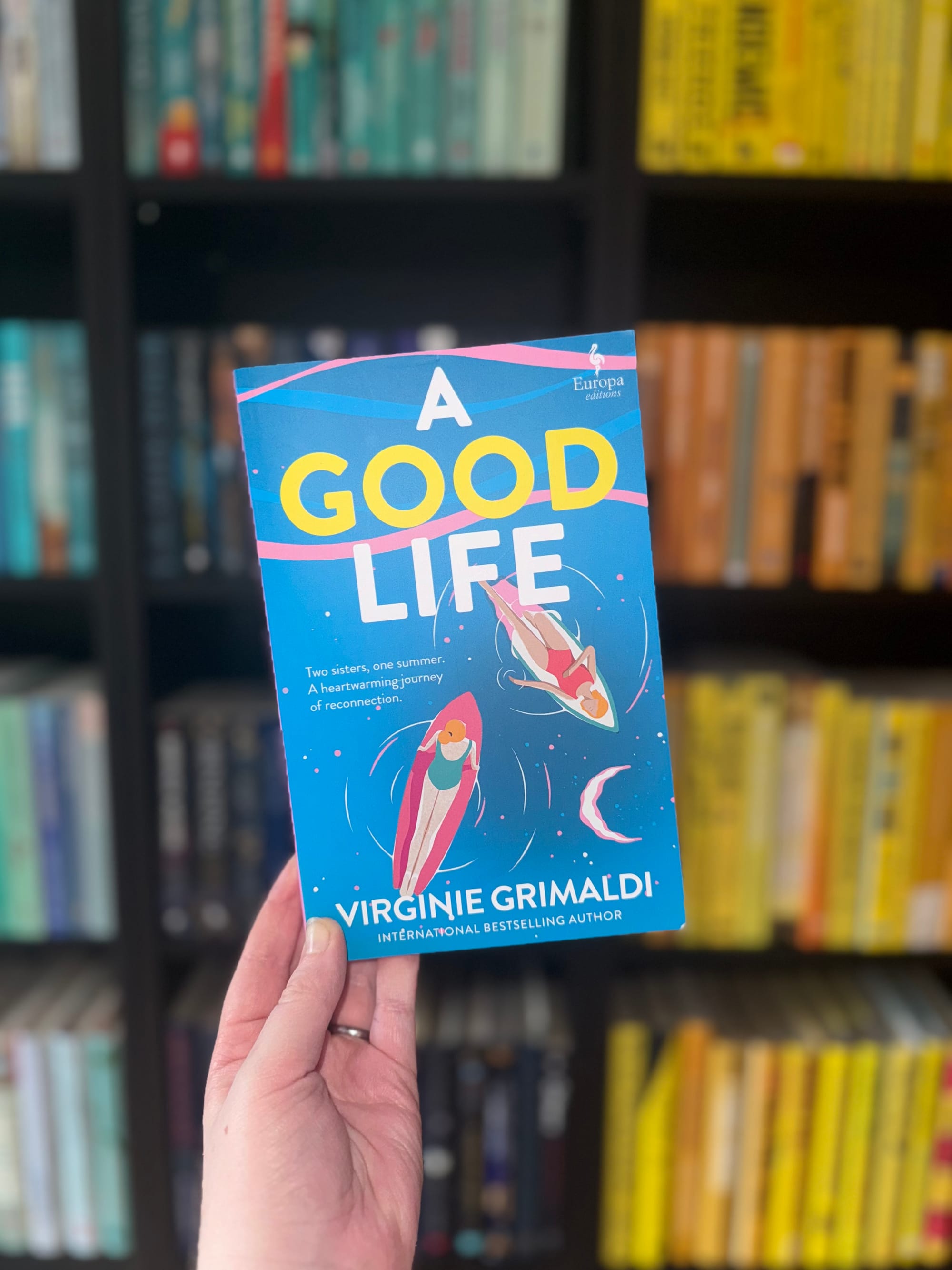 Hand holding the book "A Good Life" by Virginie Grimaldi in a library. Blue cover with illustrations of women on floats. Shelves in background.