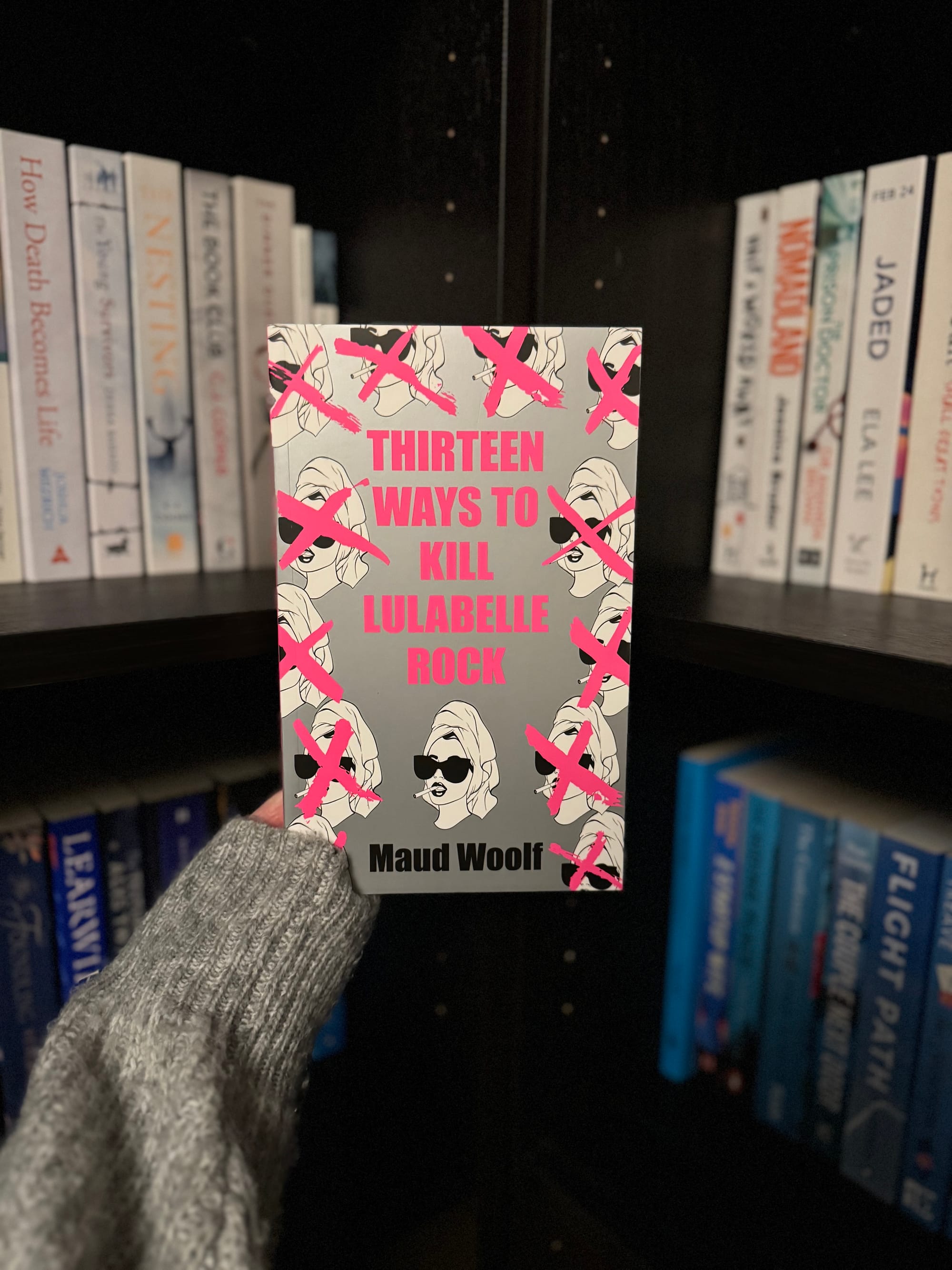 Person holds book "Thirteen Ways to Kill Lulabelle Rock" by Maud Woolf. Gray cover, pink text with crossed-out faces. Bookshelf background.