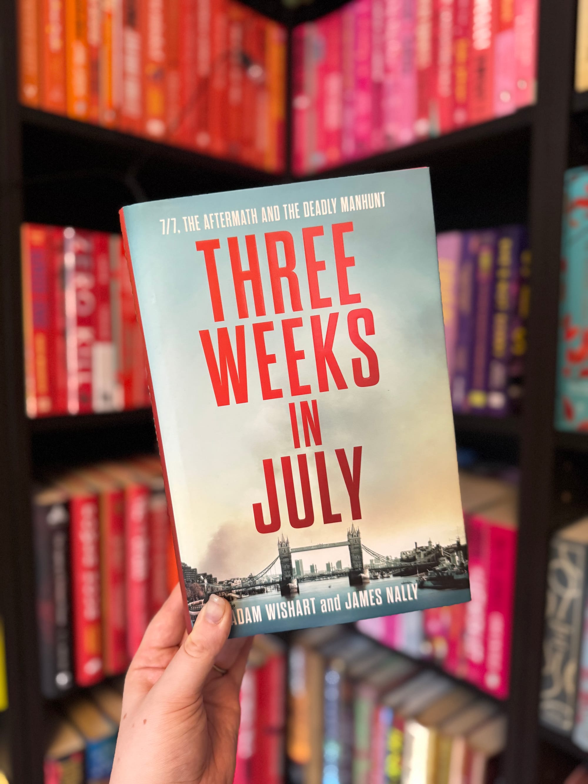 Hand holds "Three Weeks in July" book against colorful bookshelf. Red text and London Bridge image on cover. Bookstore setting.