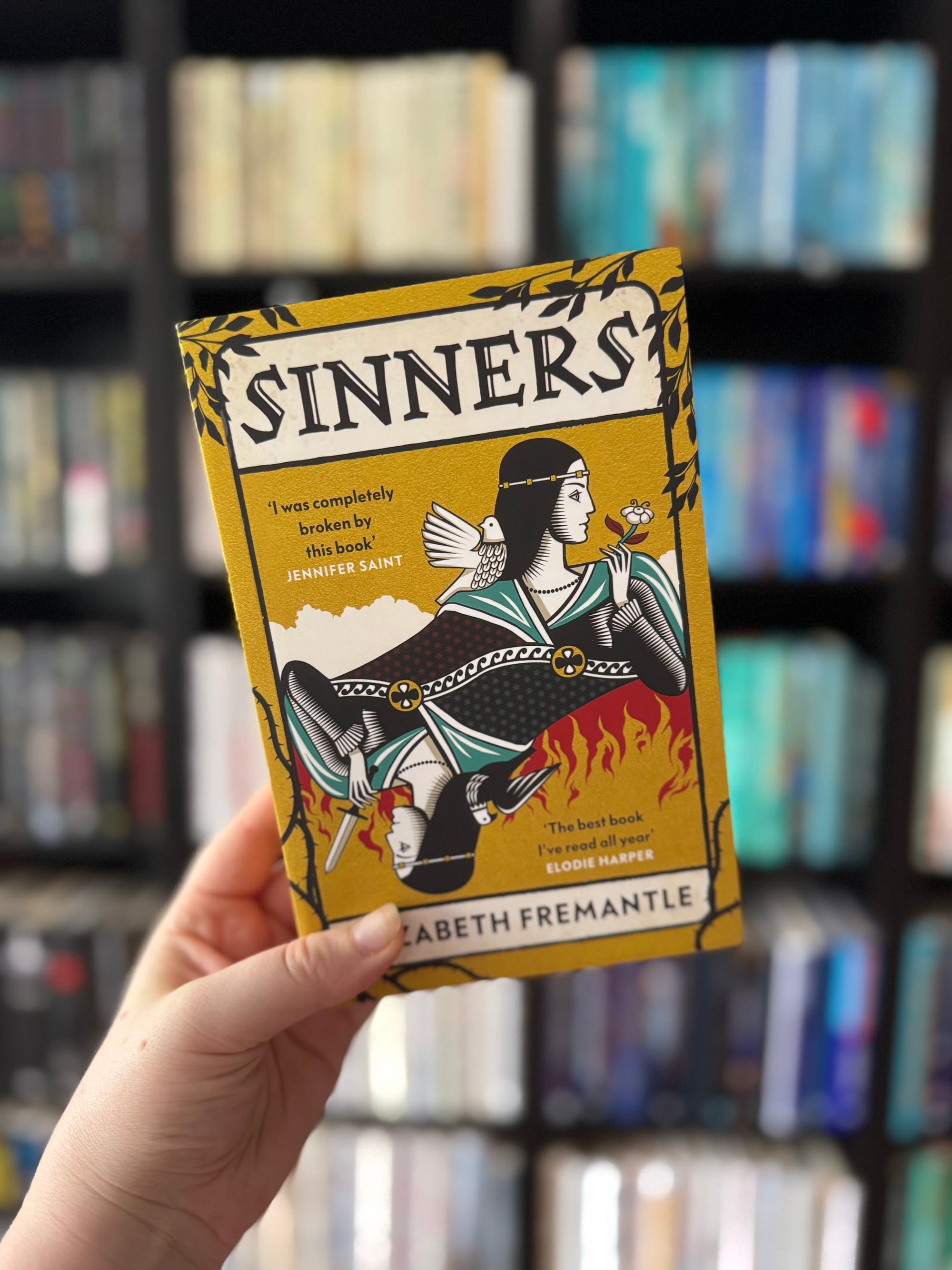 Hand holding a yellow book titled "Sinners" by Elizabeth Fremantle, with illustrations and reviews on the cover. Blurred bookshelf background.