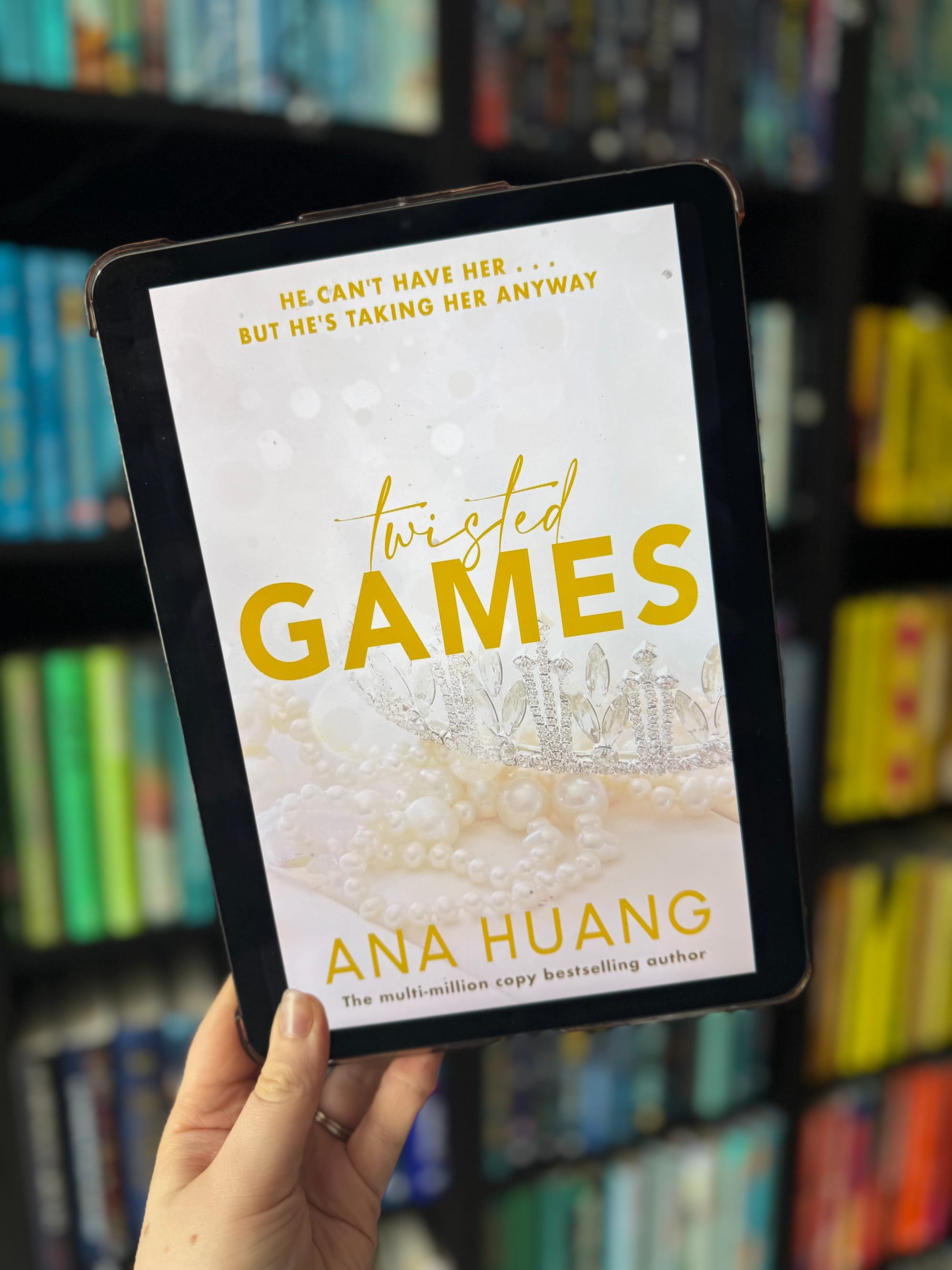 A hand holds a tablet displaying the book "Twisted Games" by Ana Huang. The cover has a crown and pearls. Blurred bookshelf background.