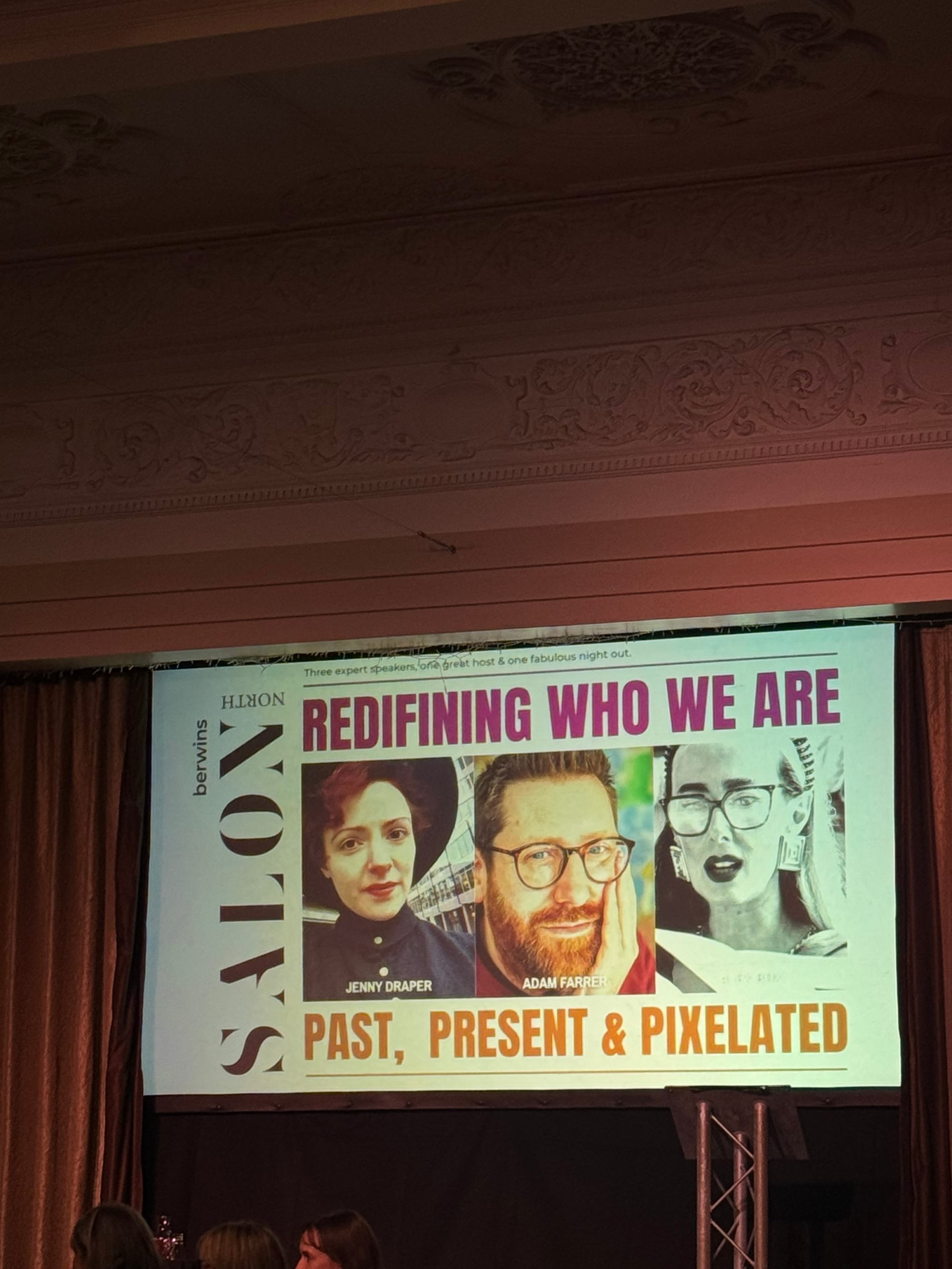 Presentation setting with a screen displaying a poster featuring three people. Text: "Redefining Who We Are. Past, Present & Pixelated."